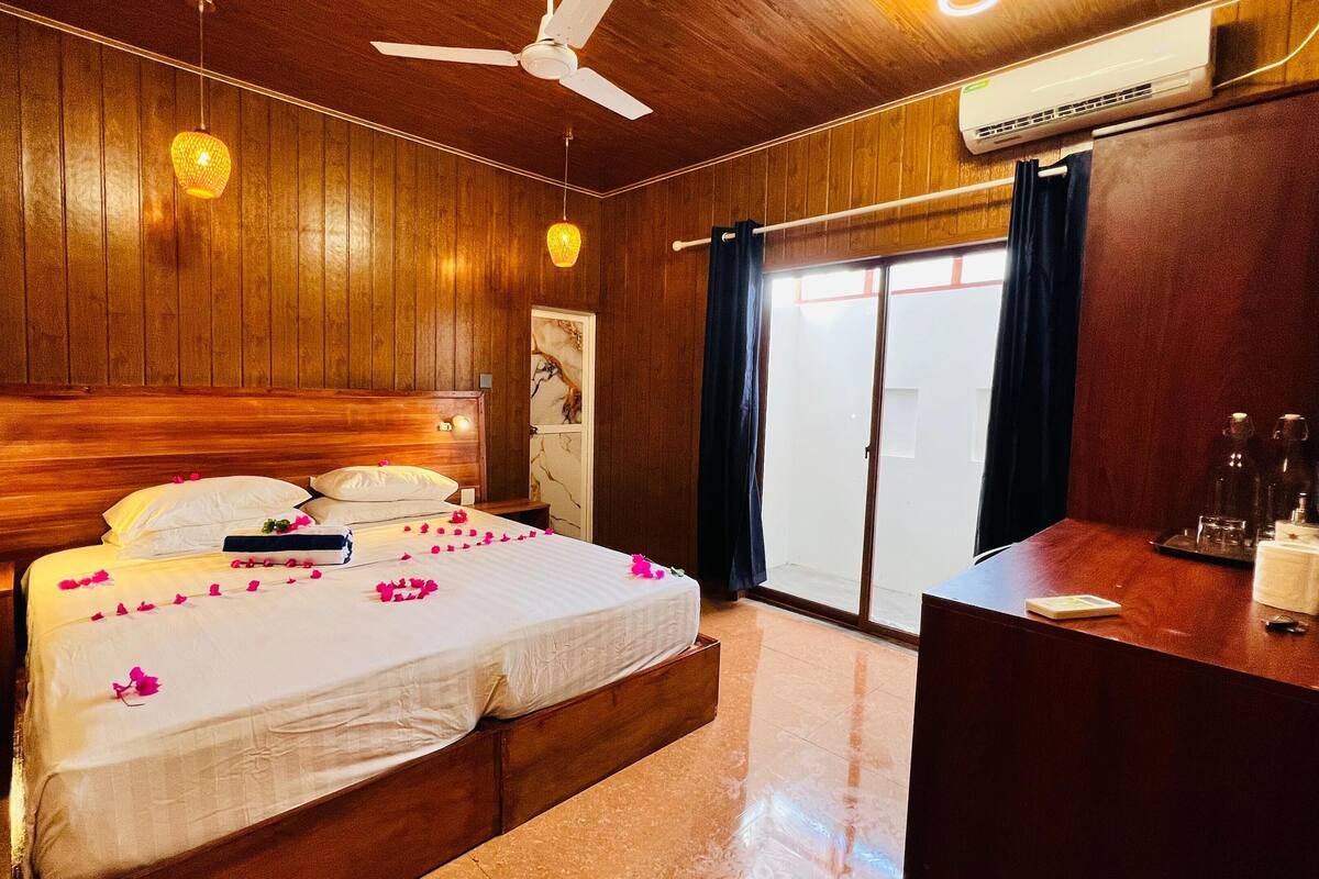 A wooden-paneled room with a bed, a desk, an air conditioner, and a ceiling fan.
