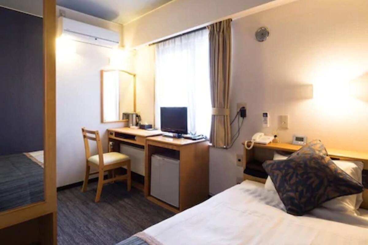 A hotel room with a bed, a desk with a computer, a chair, a small fridge, a bedside table with a lamp, and a telephone.