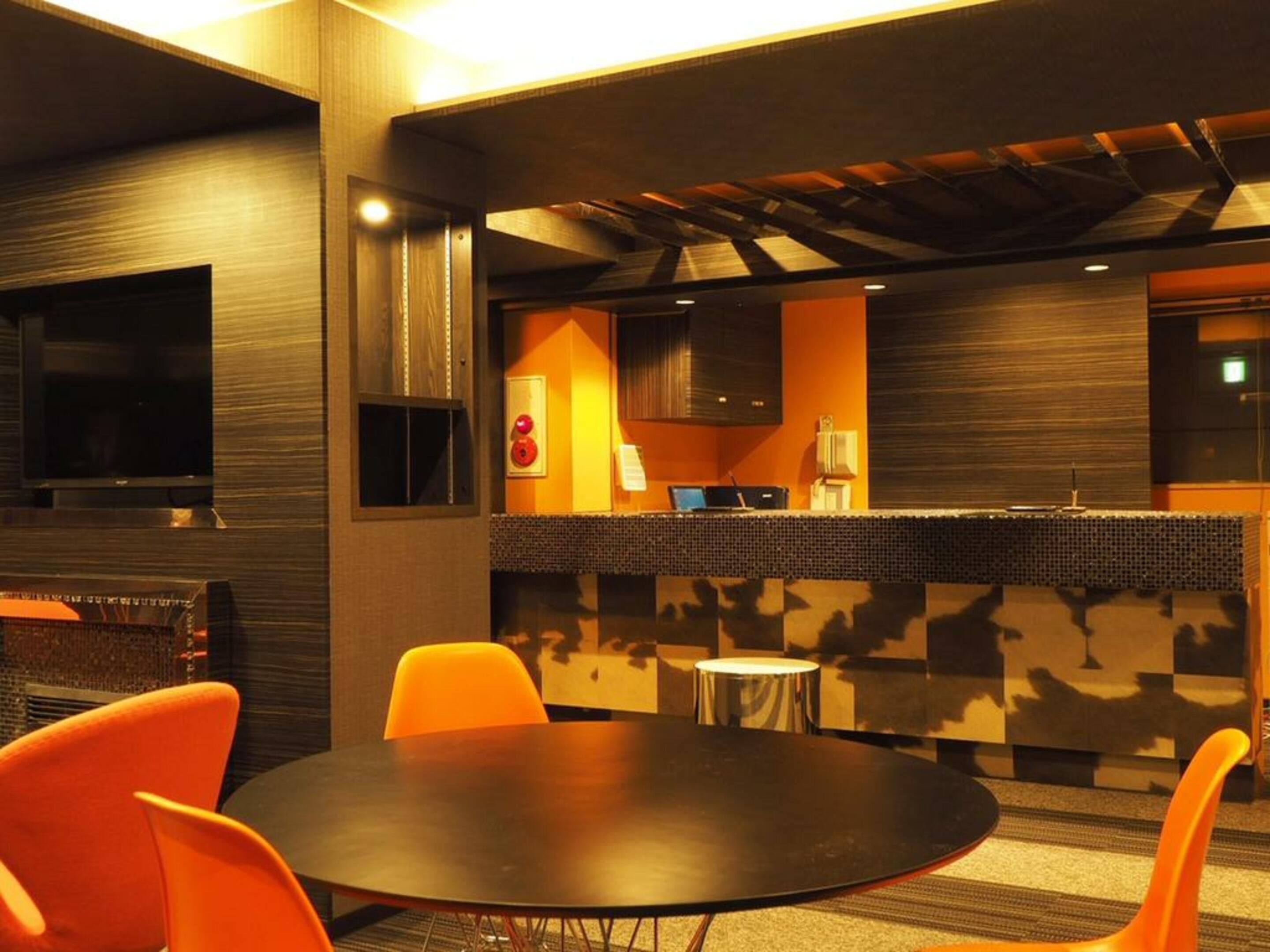 Un intérieur moderne avec une table ronde, des chaises orange, un téléviseur à écran plat, un petit coin cuisine et un mur orné d’un motif.