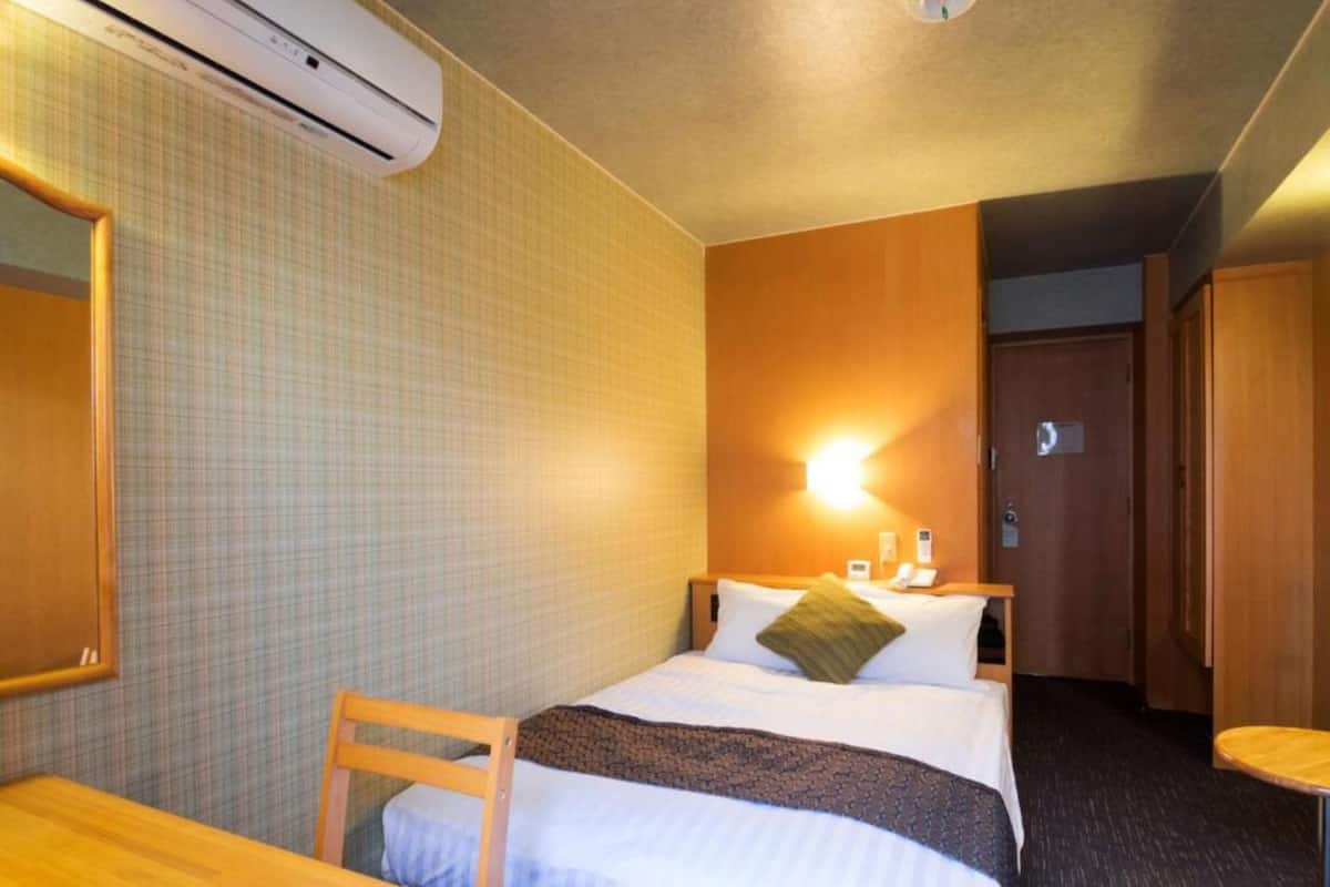 A hotel room with a bed, a desk, a chair, an air conditioning unit, a wall-mounted light, and a door.