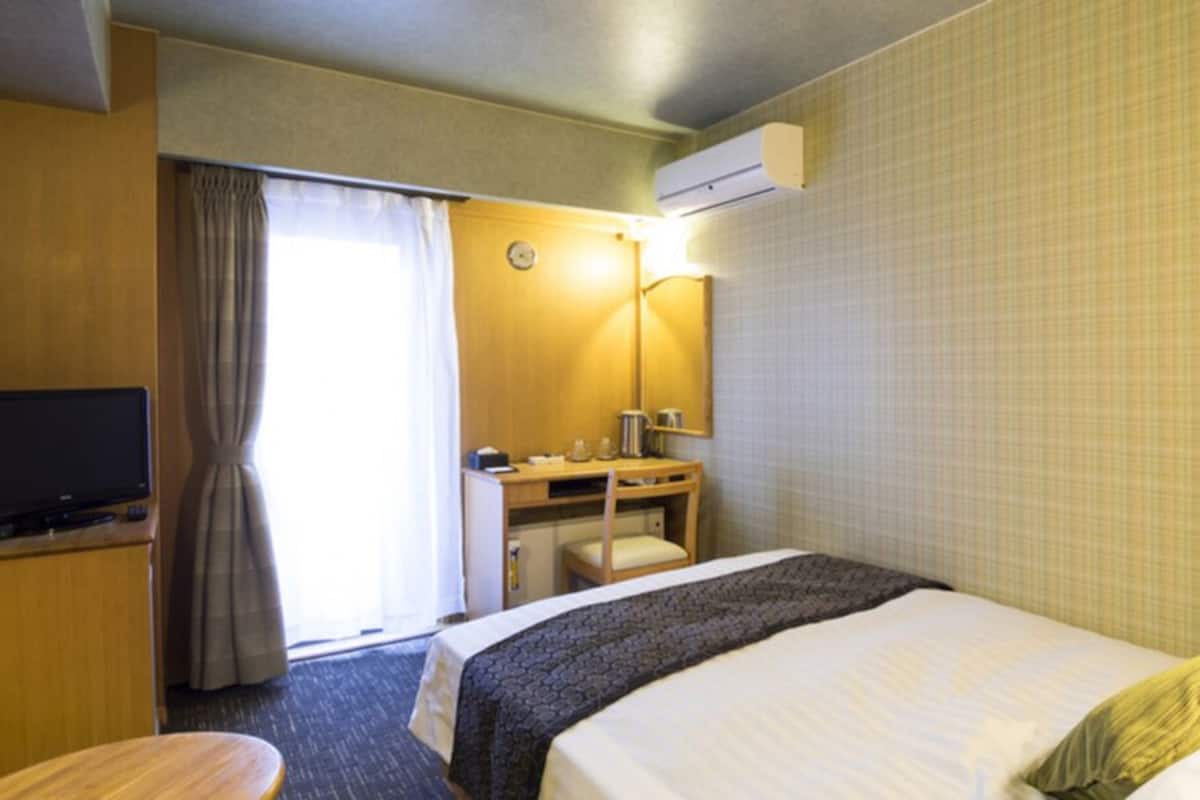 A hotel room with a bed, a desk, a chair, a television, a window with curtains, and an air conditioning unit.