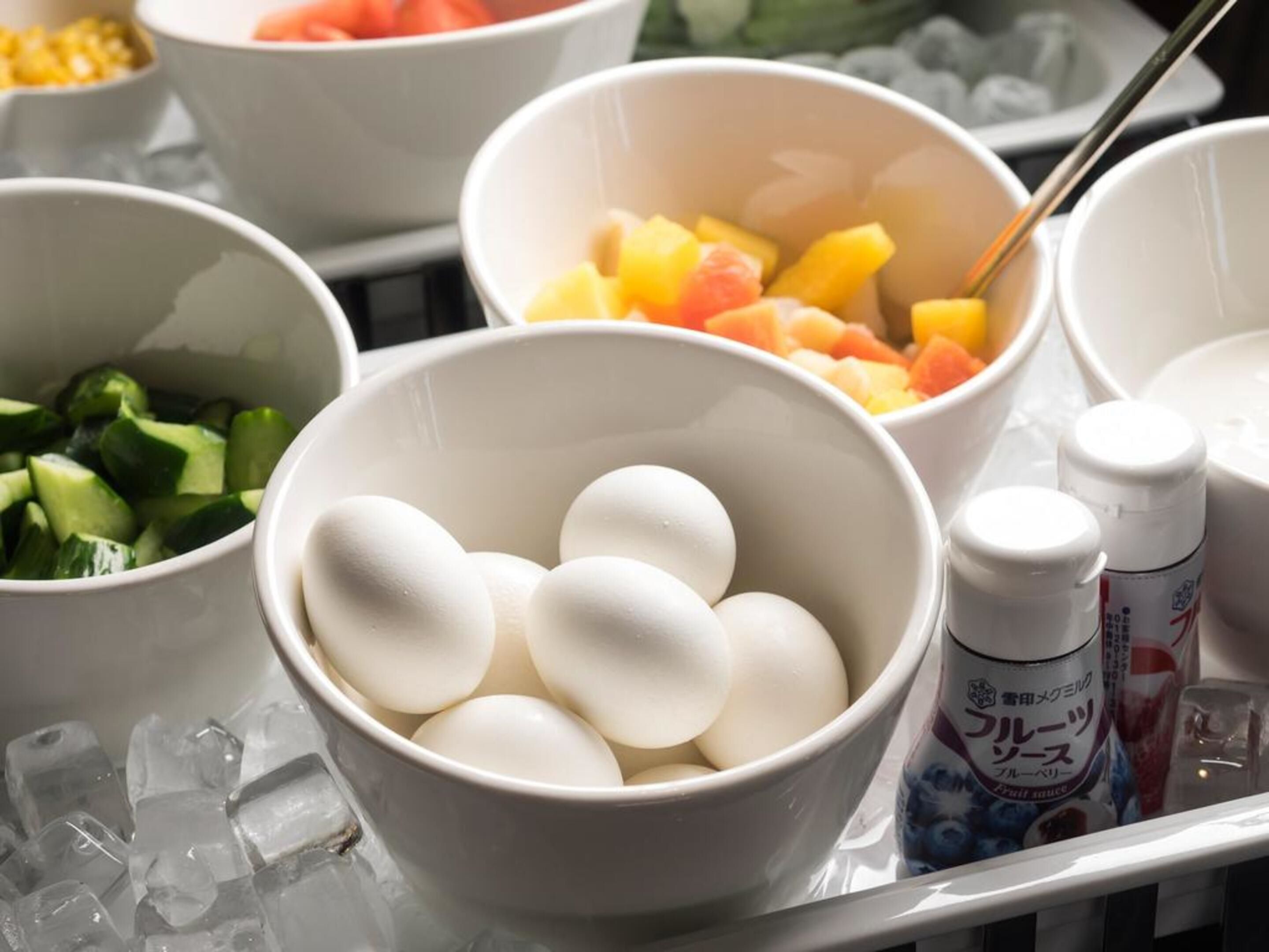 A tray with bowls containing eggs, diced vegetables, and fruit, along with condiments and ice cubes.