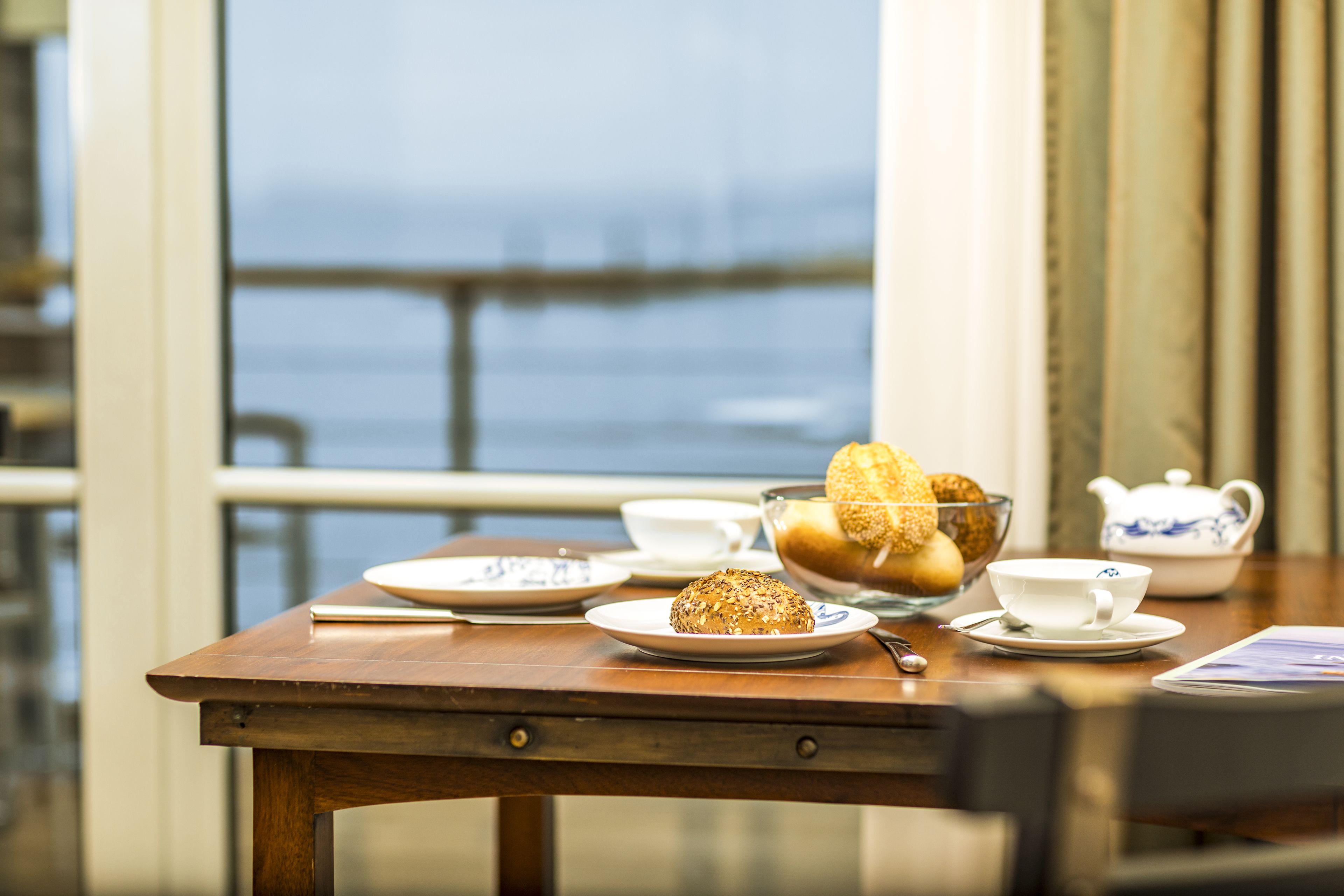 A table with a bowl of bread, a teapot, and a cup, set against a window with a view of the sea.