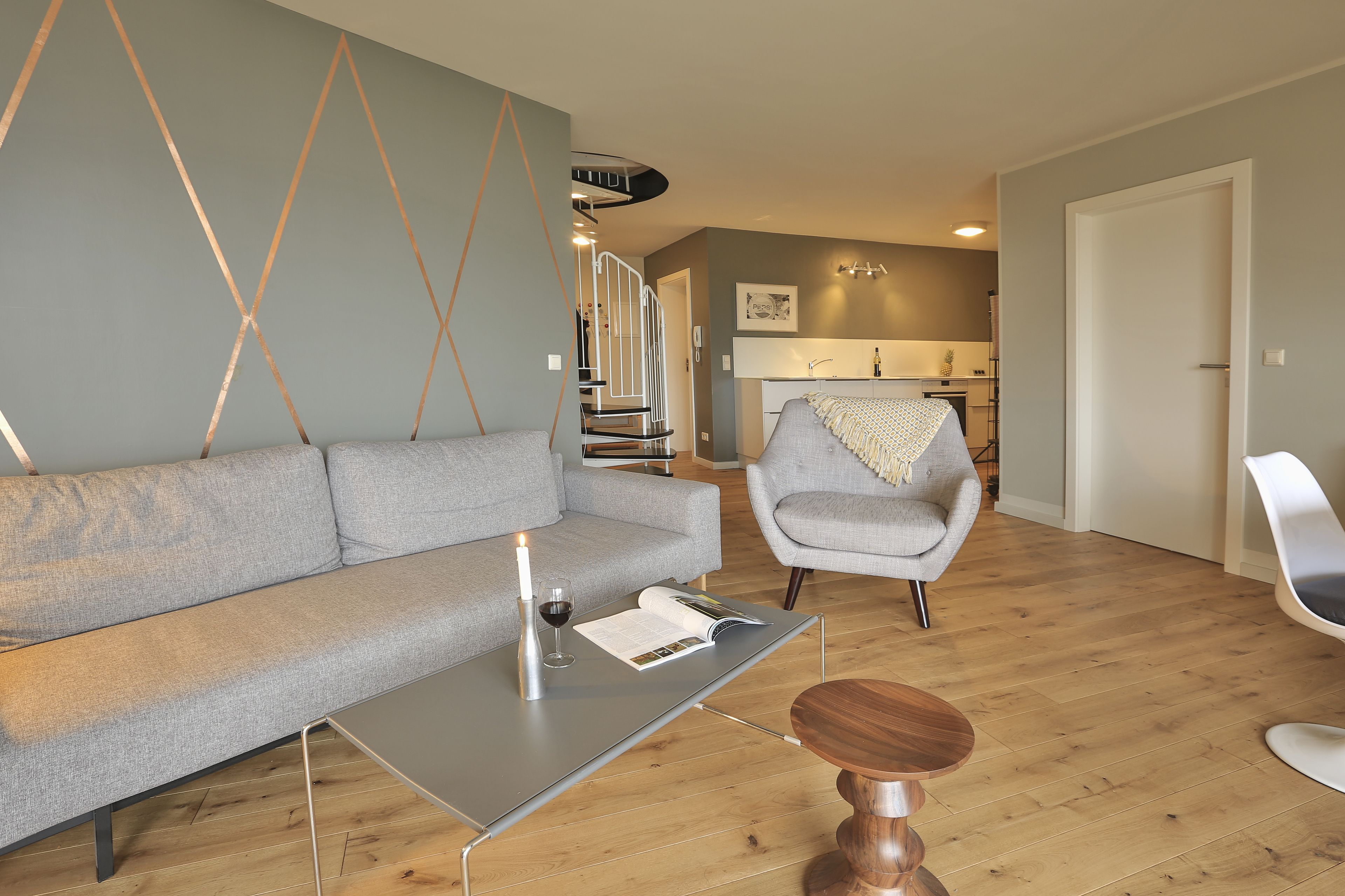 A modern living room with a grey sofa, a coffee table, and a wooden chair. A staircase leads to an upper level.