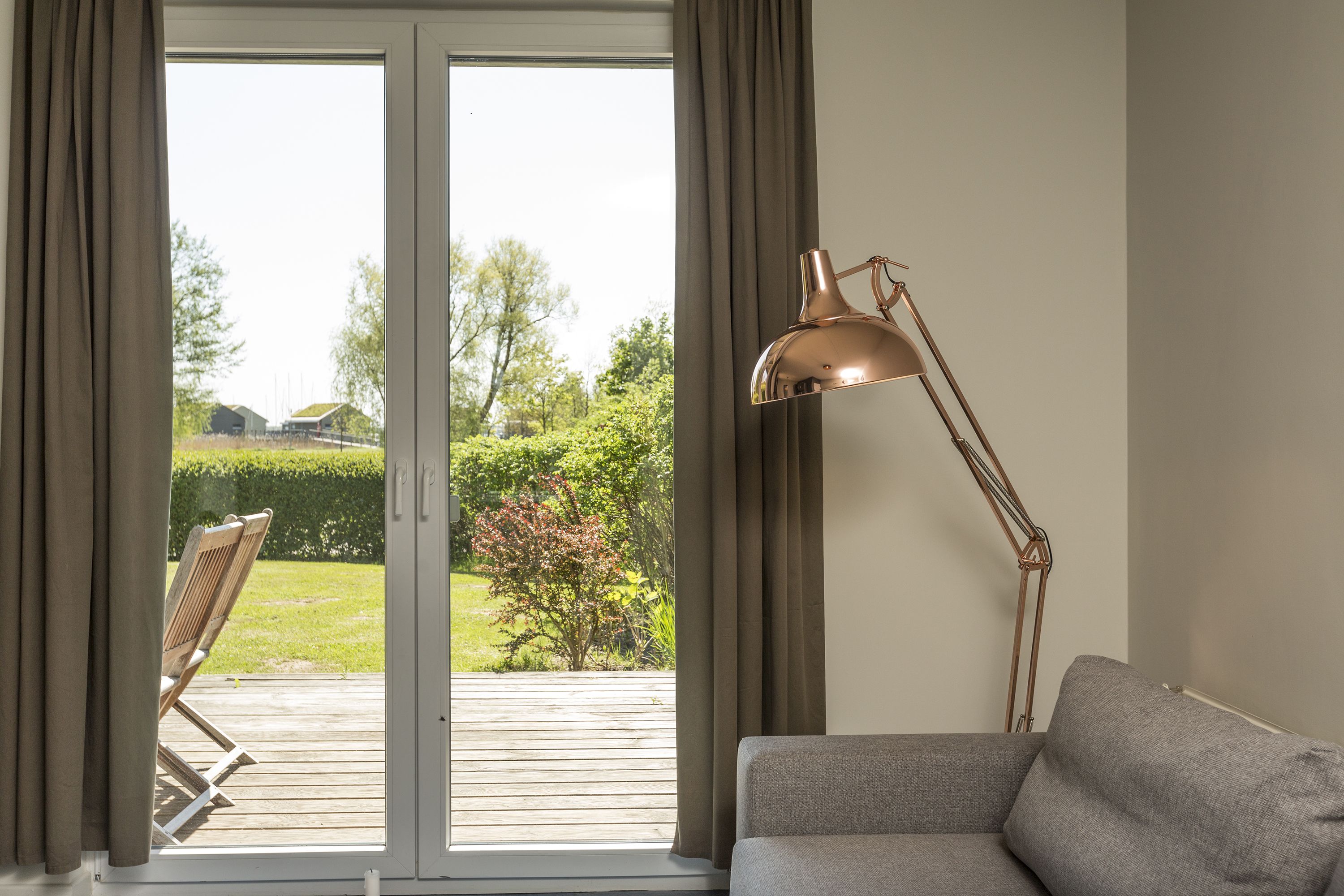 Een kamer met een grijze bank, een vloerlamp en een uitzicht door een schuifdeur op een terras.