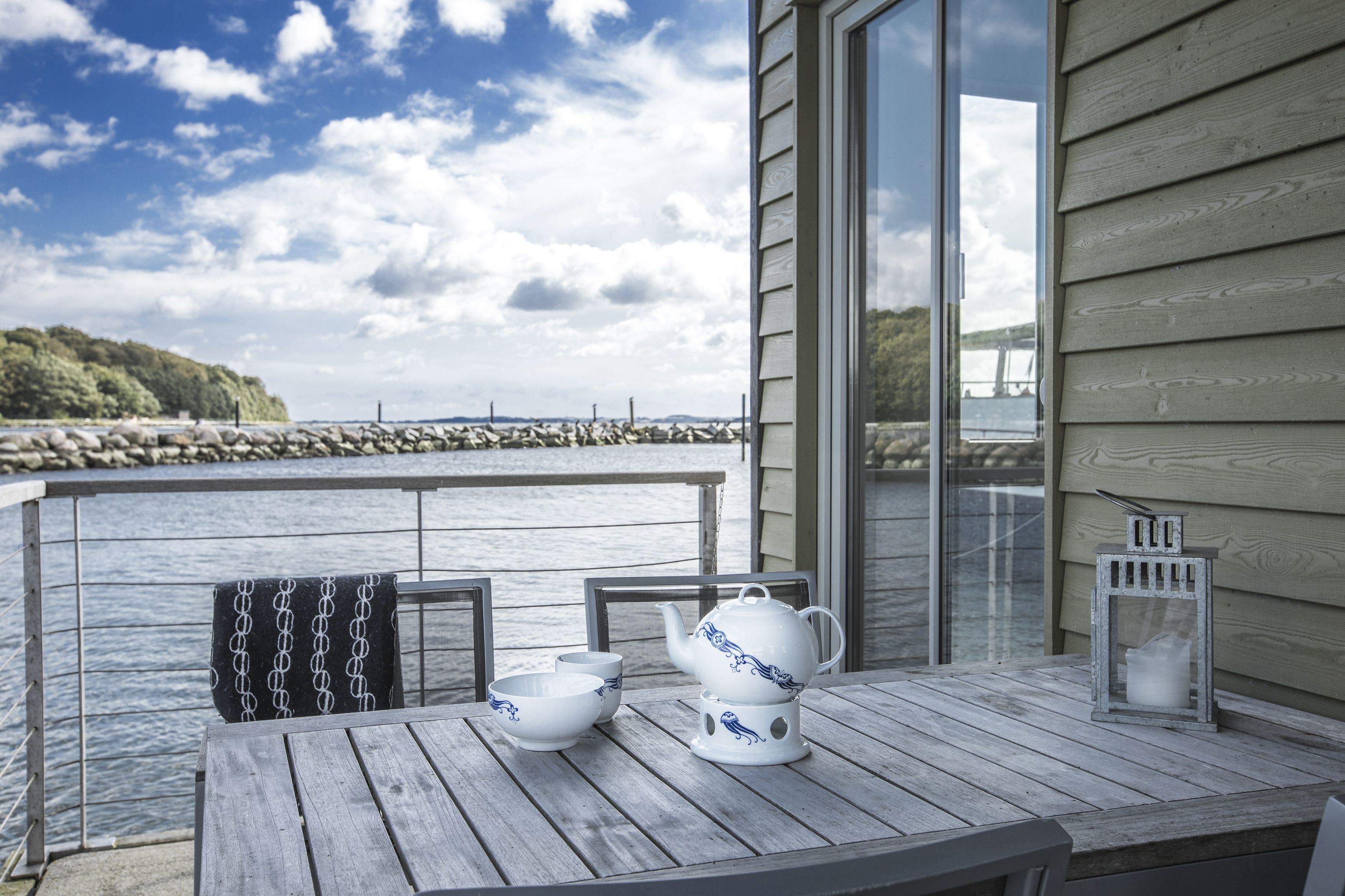 Een houten terras met een gedekte tafel voor thee, met uitzicht op een wateroppervlak en een rotsachtige kustlijn.