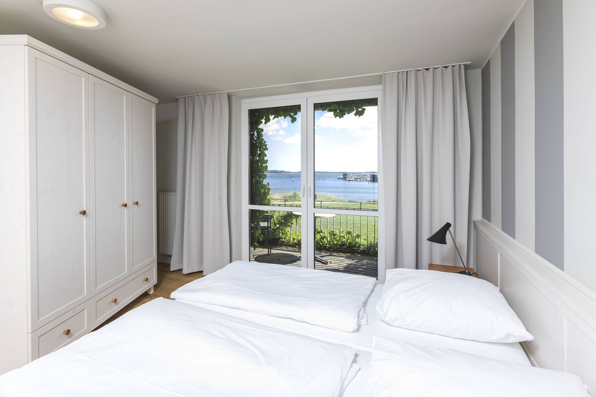 A bedroom with two beds, a large wardrobe, and a view of a body of water through a glass door.