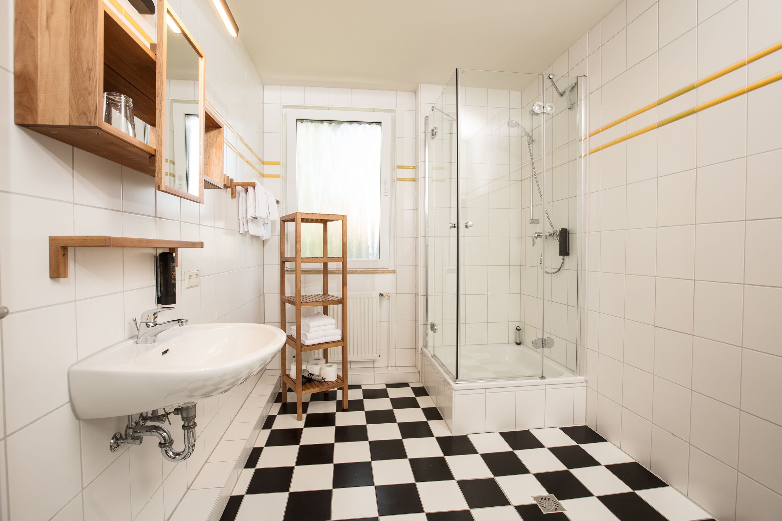 Une salle de bain avec un lavabo blanc, des étagères en bois, une cabine de douche en verre et un carrelage à carreaux.