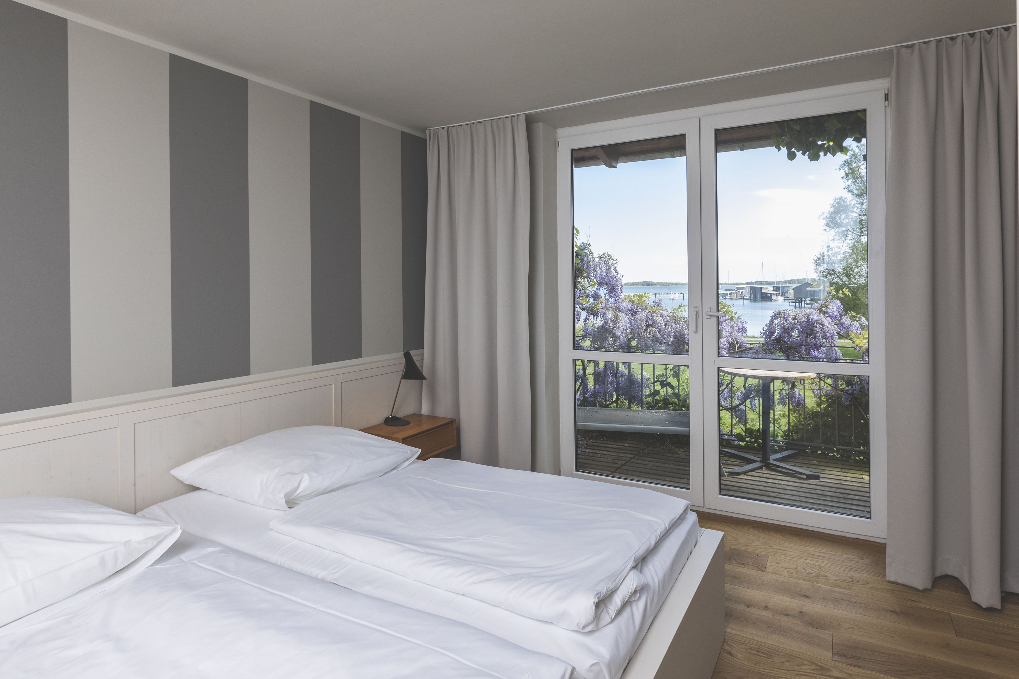 A bedroom with two beds, a nightstand, a window with a view of the outdoors, and a balcony with purple flowers.
