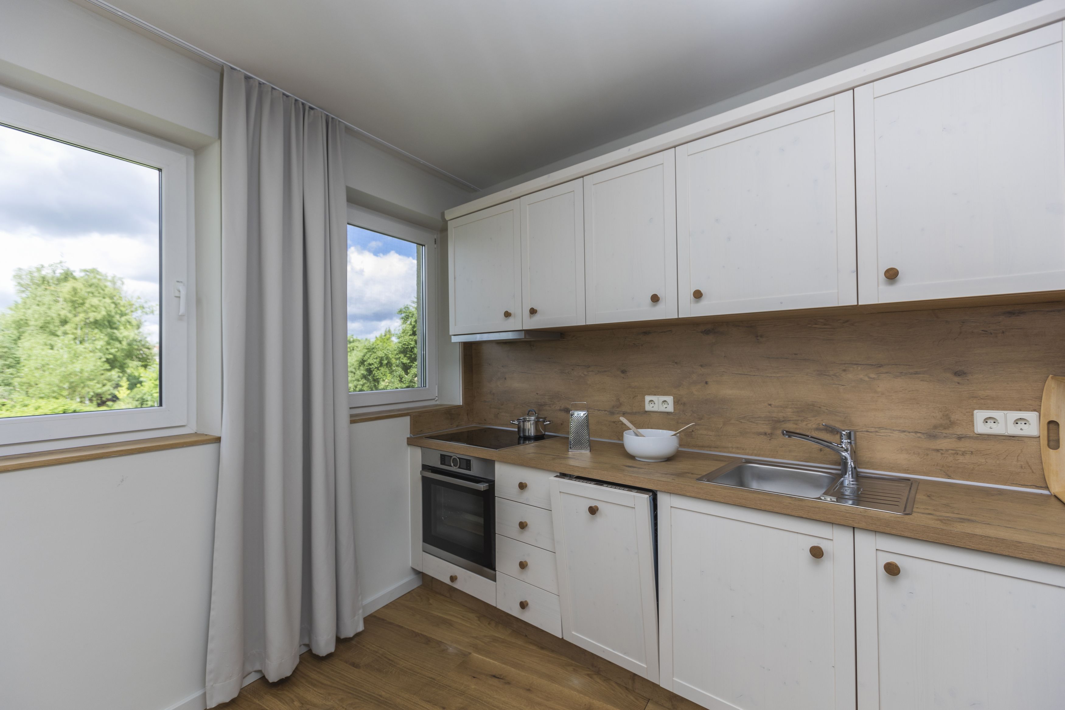 Una cocina con armarios blancos, encimera de madera, horno empotrado y ventana con cortinas.
