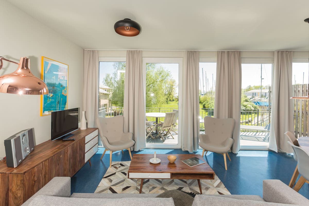 A modern living room with a wooden coffee table, two armchairs, a TV, and a large window with a view of a garden and pool.