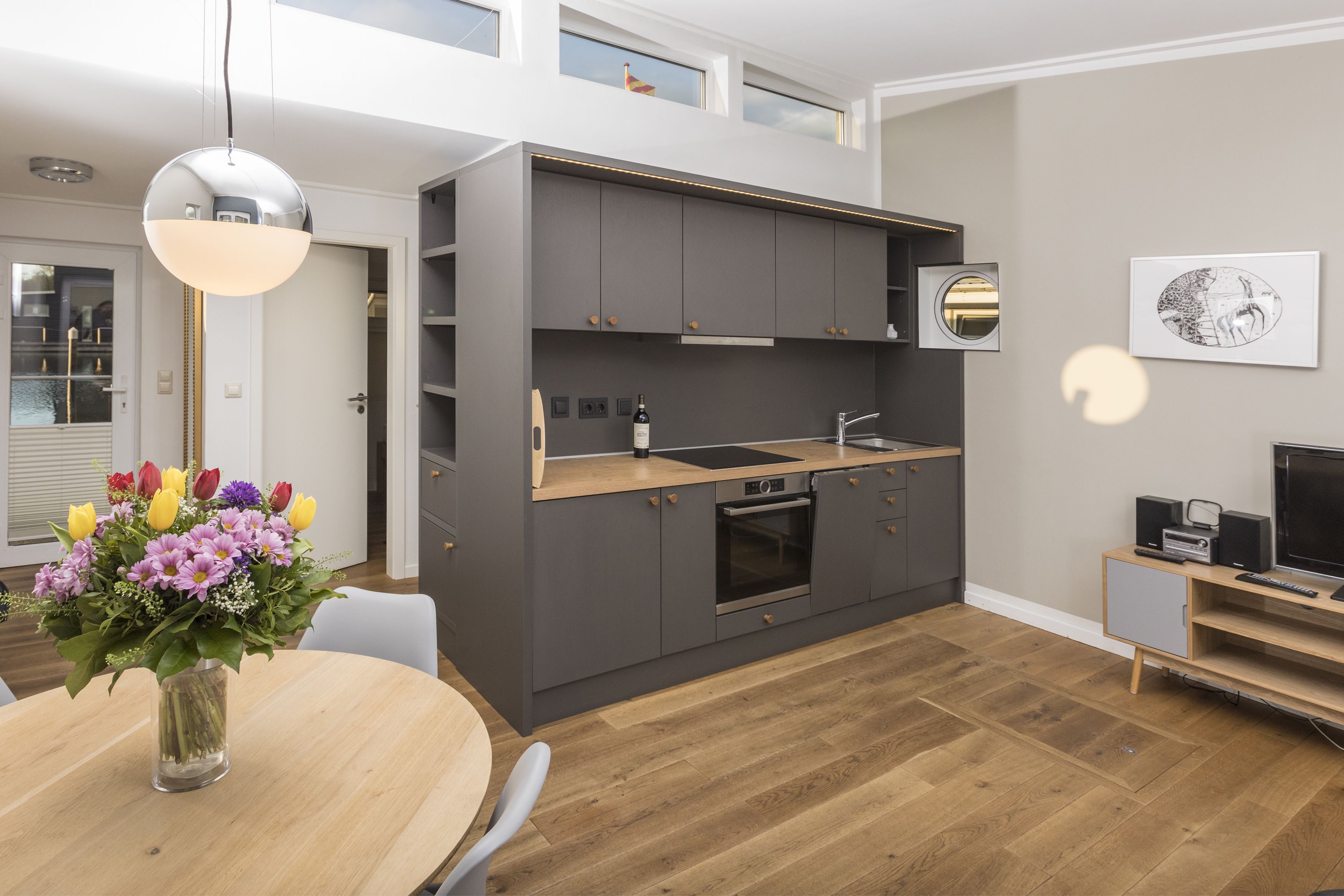 Une cuisine moderne avec des meubles gris, un plan de travail en bois et un four encastré. On y trouve une table à manger, un vase de fleurs et une chaise.
