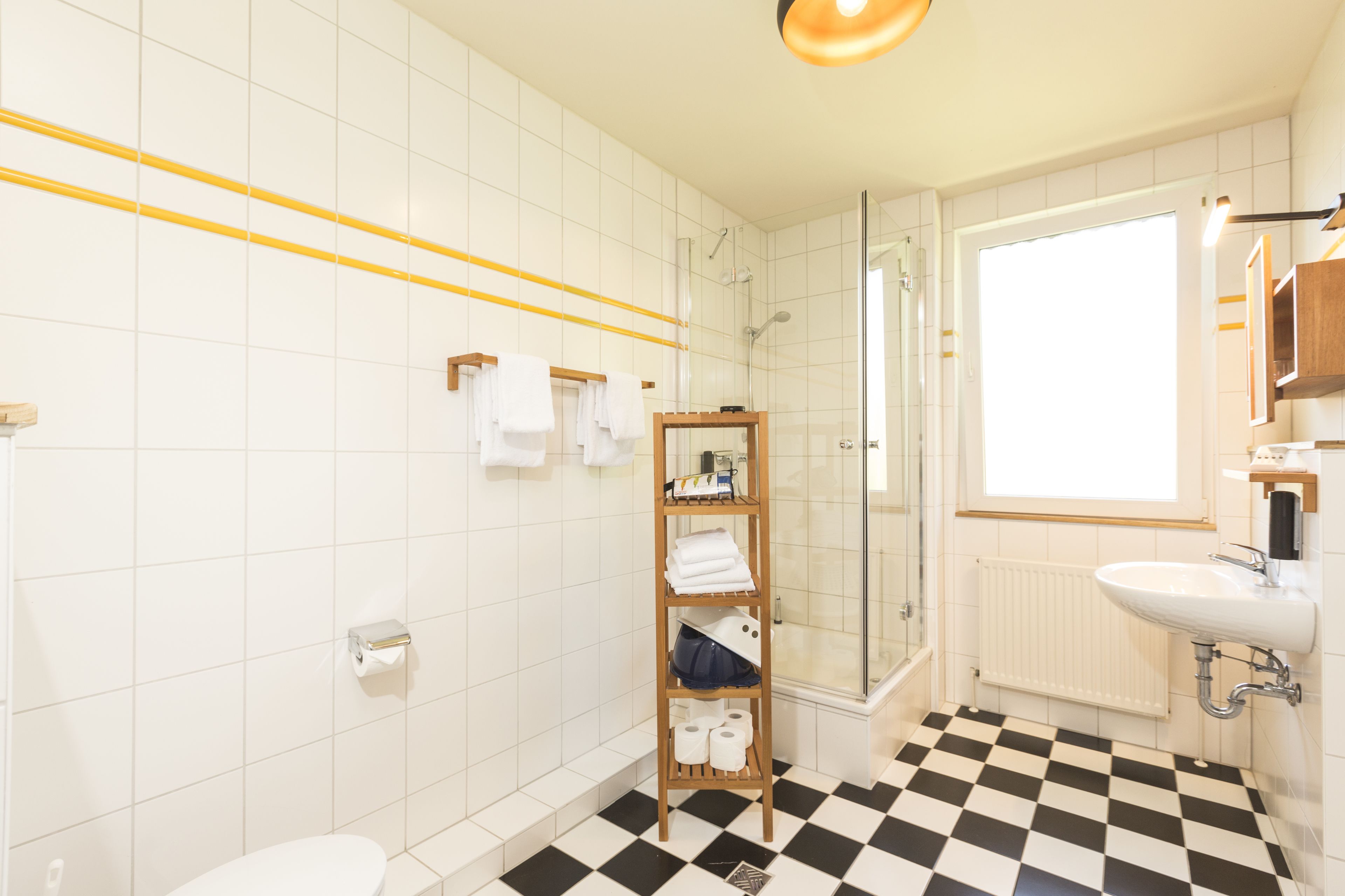 Un baño con piso a cuadros blancos y negros, un estante de madera con toallas, un box de ducha de vidrio y un lavamanos blanco con espejo superior.