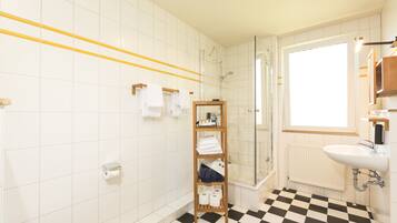 Un baño con piso a cuadros blancos y negros, un estante de madera con toallas, un box de ducha de vidrio y un lavamanos blanco con espejo superior.