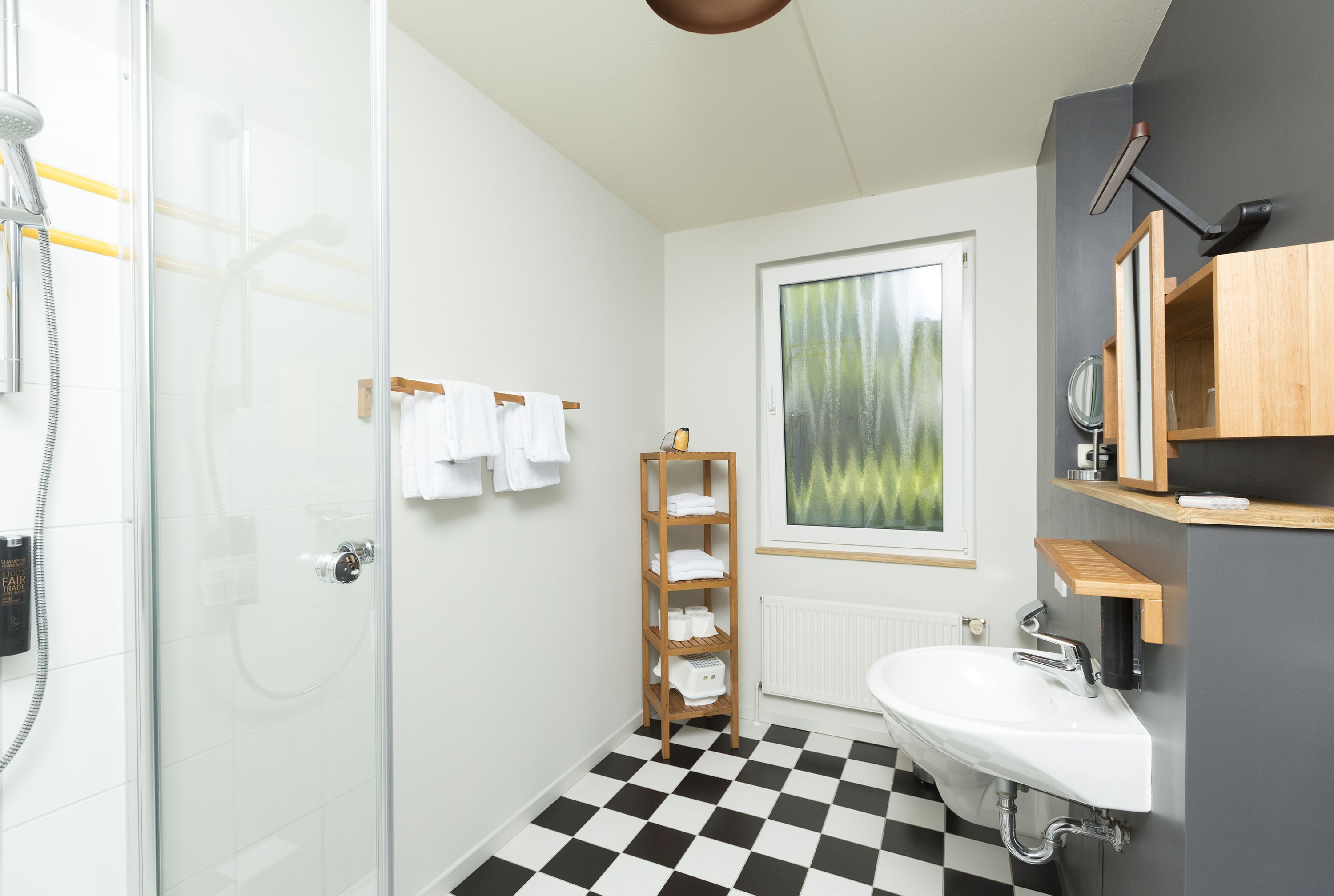 Un baño con cabina de ducha de vidrio, suelo a cuadros, lavamanos blanco y repisa de madera con toallas.