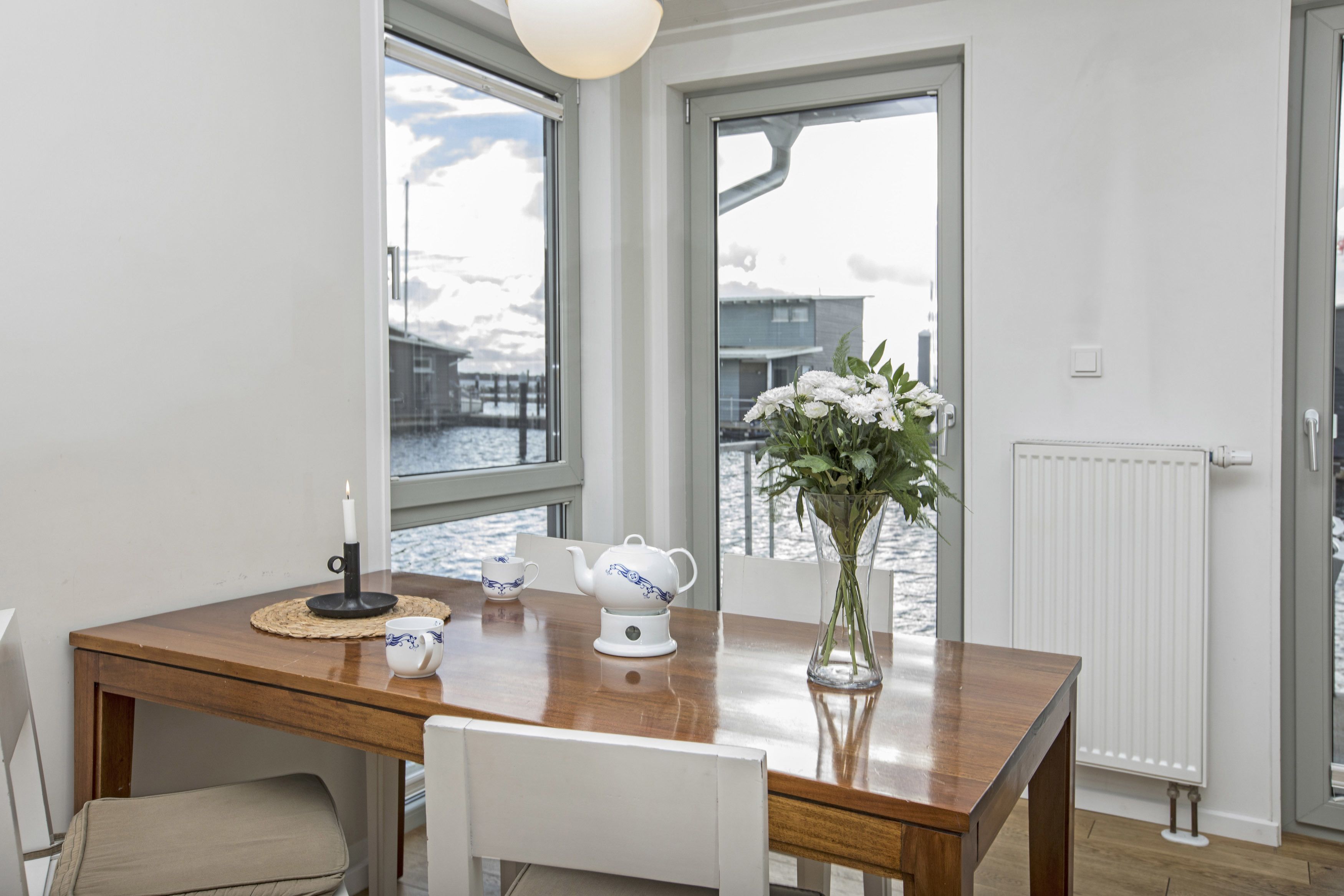 A dining area with a wooden table, white chairs, a vase with flowers, a white teapot, and a candle.