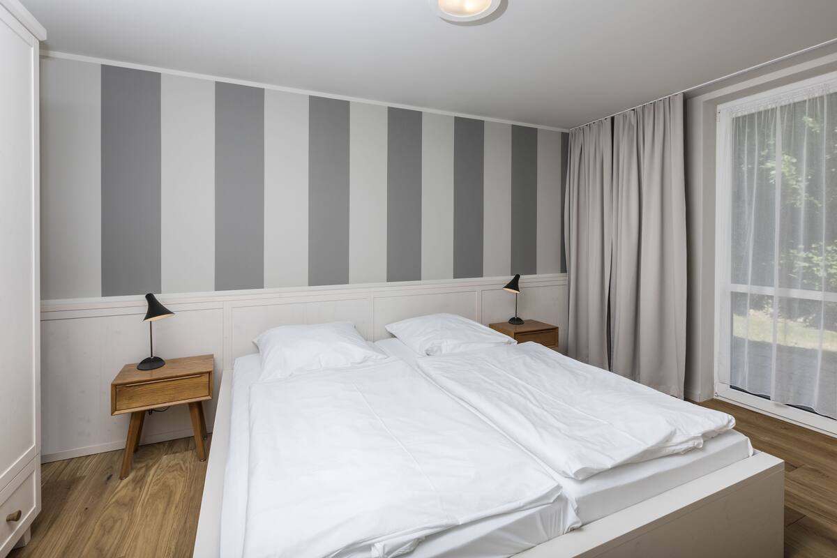 A double bed with white bedding, two bedside tables with lamps, a striped wall, and a window with curtains.