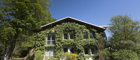 Una casa de dos plantas con techo inclinado, rodeada de abundante vegetación y árboles.