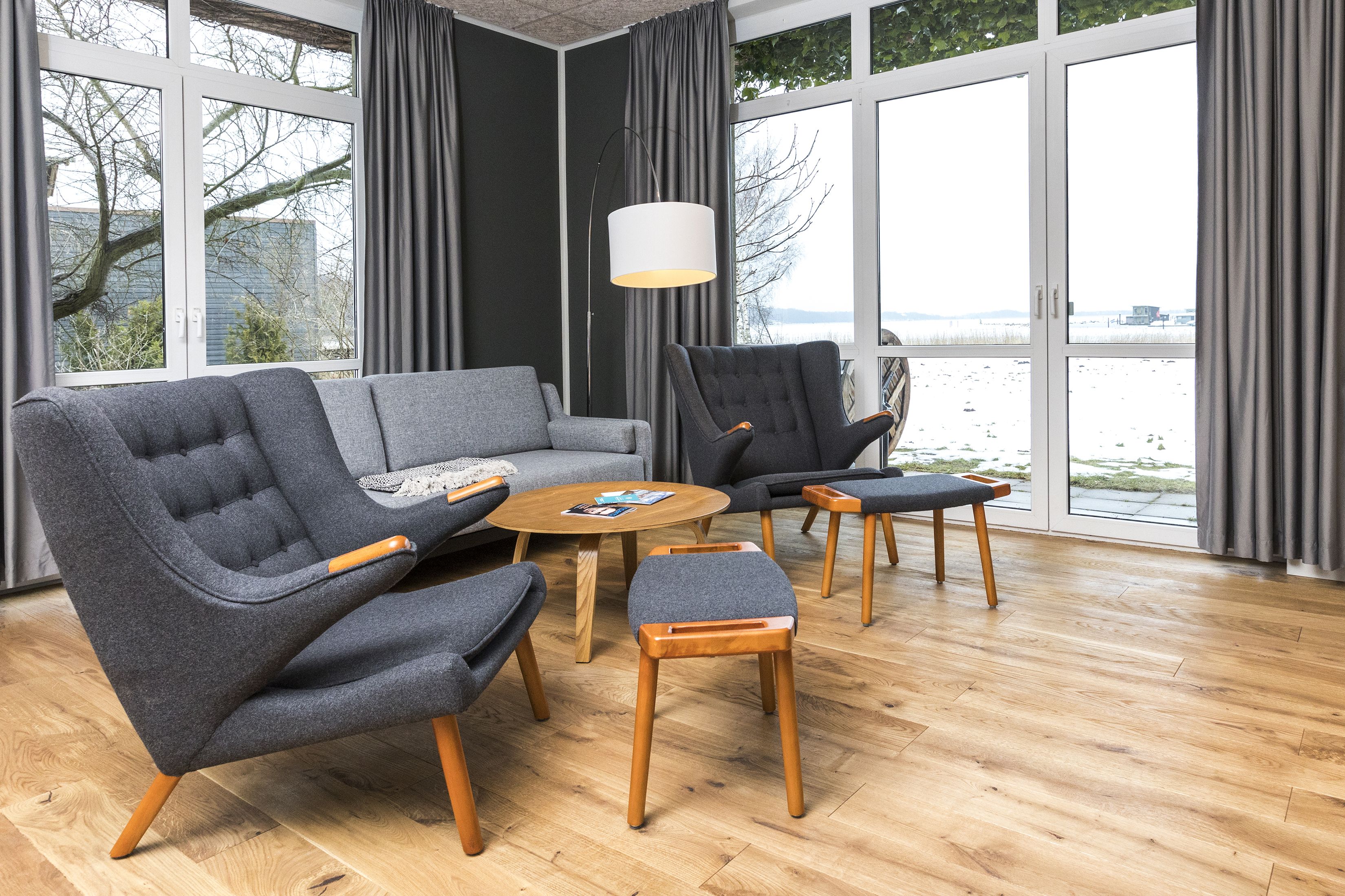 A modern living room with a grey sofa, two armchairs, a coffee table, and a wooden floor. Large windows offer a view of the outdoors.