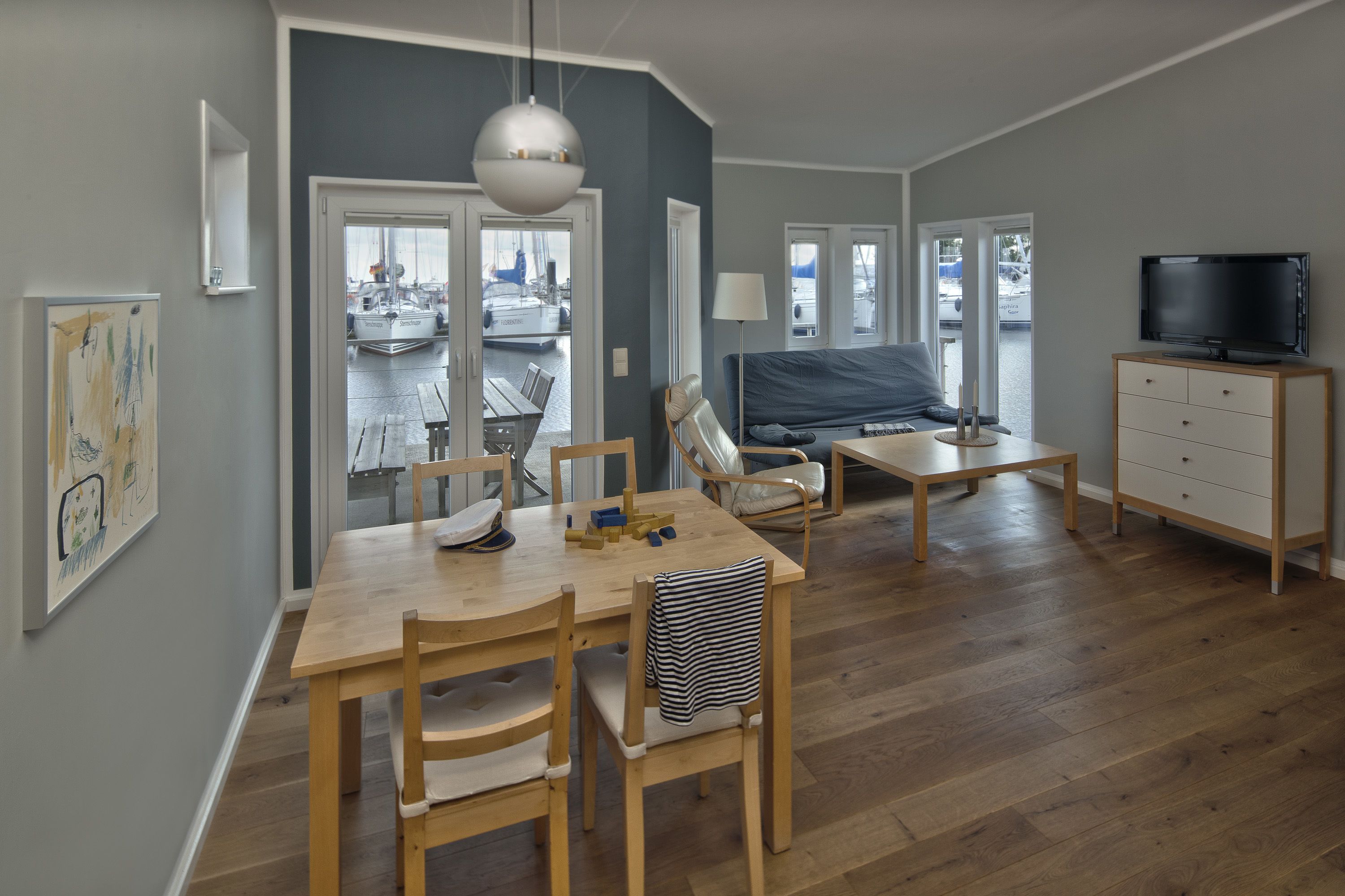 A modern living room with a wooden dining table, chairs, a sofa, a TV, and a view of the outside.