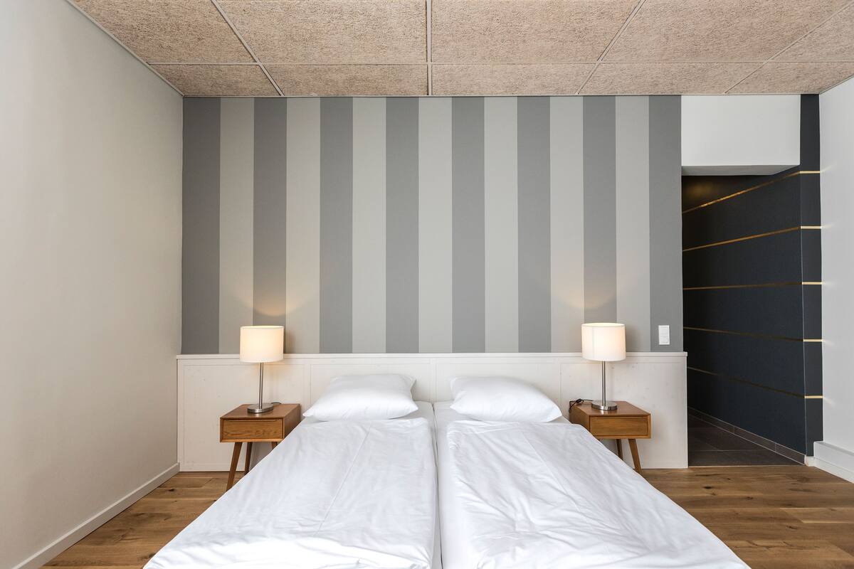 A double bed with white linens, two bedside tables with lamps, a striped wall, and a staircase with a dark wall and gold lines.