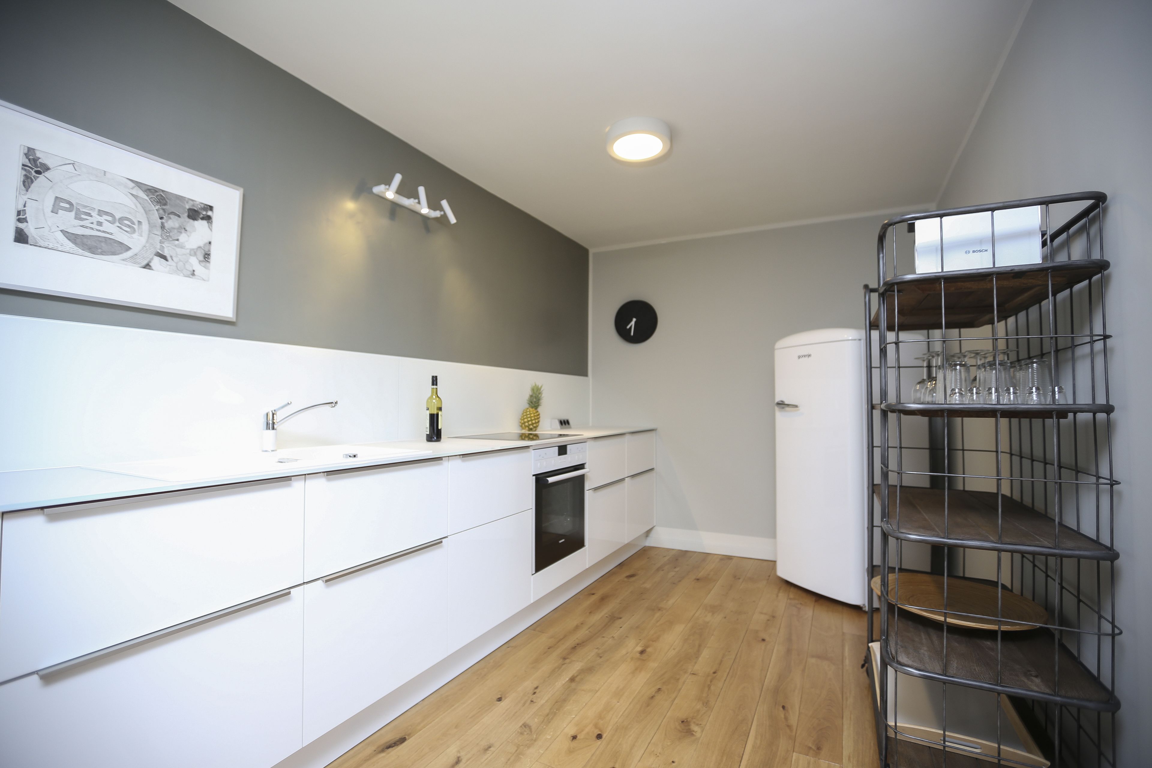 A modern kitchen with white cabinets, a stainless steel sink, a built-in oven, a white refrigerator, and a metal shelf unit with glasses.