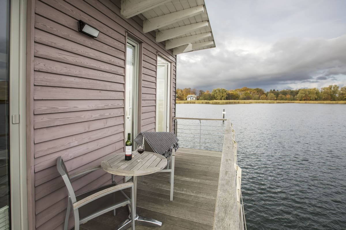 A wooden deck with a table and chairs, overlooking a body of water and a distant house.