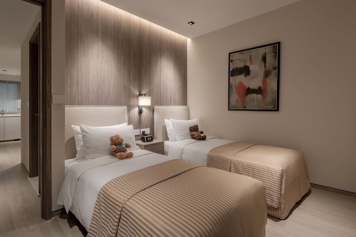 Una habitación de hotel moderna con dos camas, una mesita de noche con lámpara, un osito de peluche en una cama y un cuadro en la pared.
