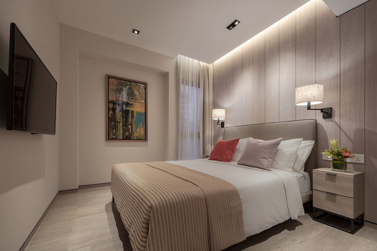 Una habitación de hotel moderna con una cama grande, un televisor de pantalla plana, un cuadro en la pared y una mesita de noche con una lámpara y un jarrón de flores.