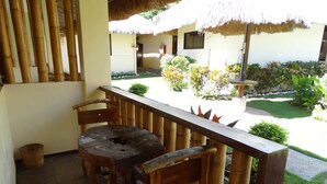 Ein Holztisch und -stühle auf einem Balkon mit einem Reetdach, mit Blick auf einen Garten mit verschiedenen Pflanzen und ein weißes Gebäude im Hintergrund.