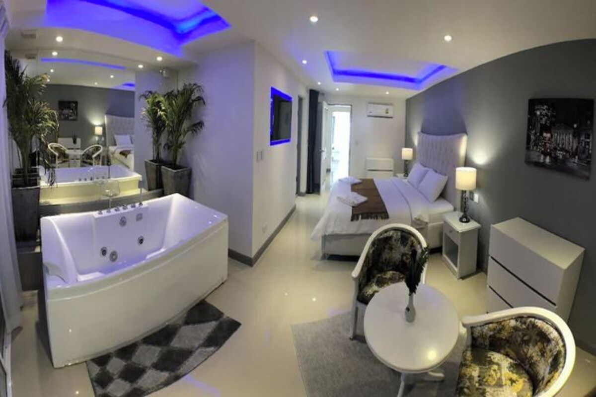 A modern bedroom with a large bathtub, a bed, a bedside table, and a chair.