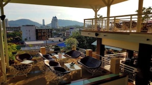 Una terraza en la azotea con mobiliario de mimbre y una zona de bar, con vistas a un paisaje urbano con montañas al fondo.