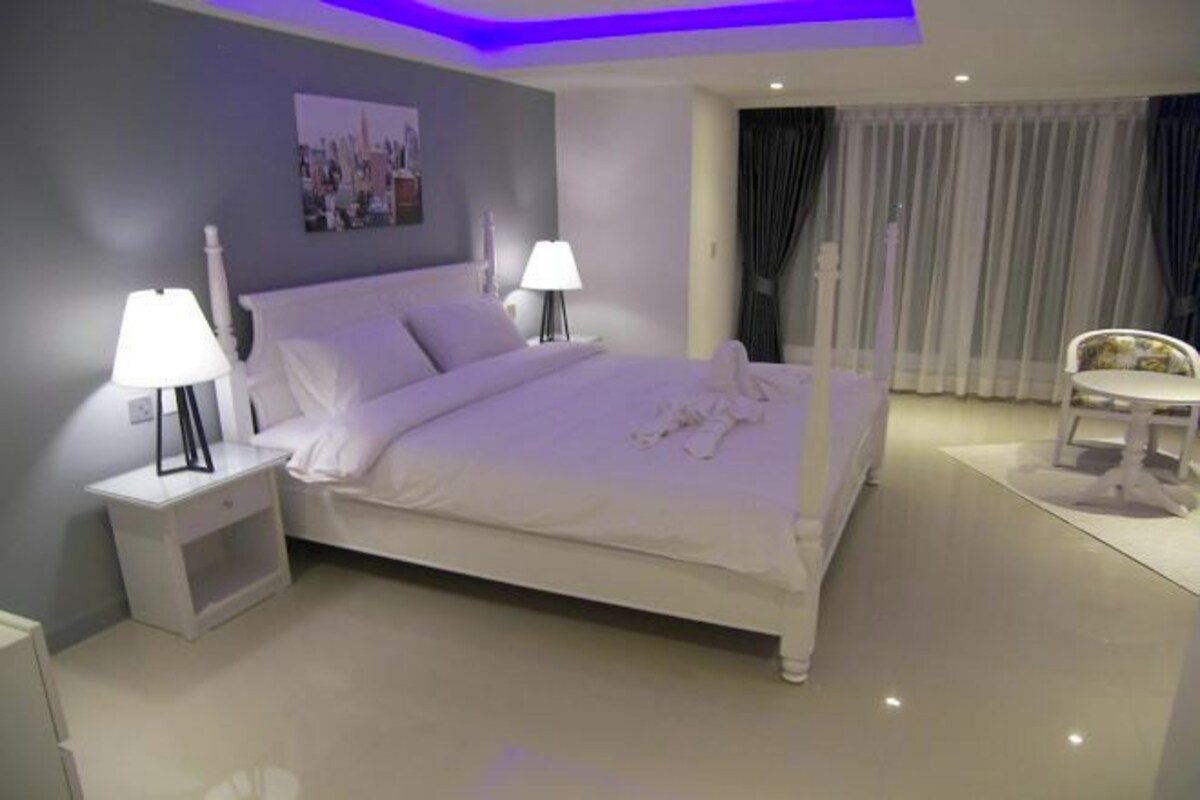 Un dormitorio moderno con una cama grande, dos mesitas de noche con lámparas, una silla y vistas a la ciudad desde la ventana.