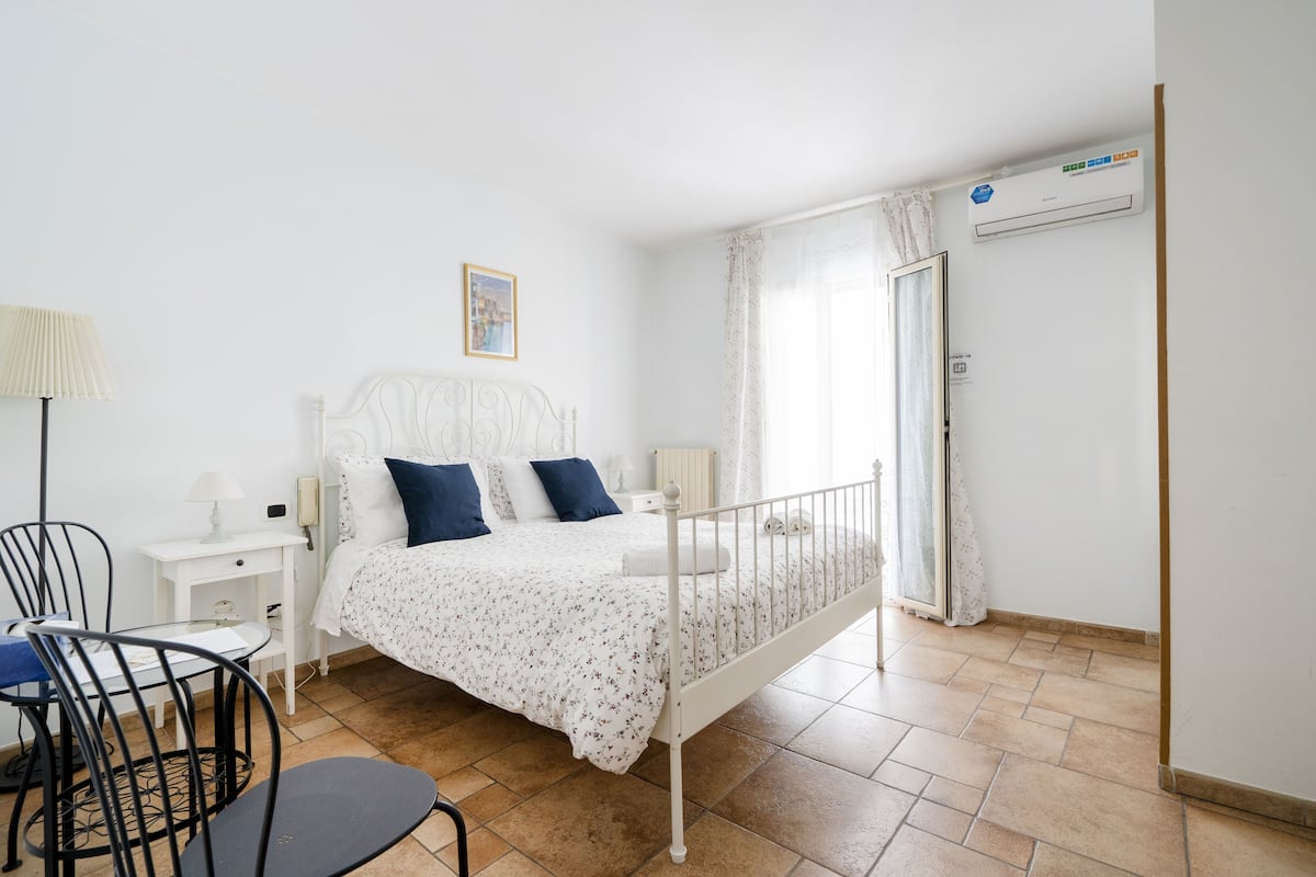 A bedroom with a bed, a chair, a nightstand, a lamp, and an air conditioning unit.