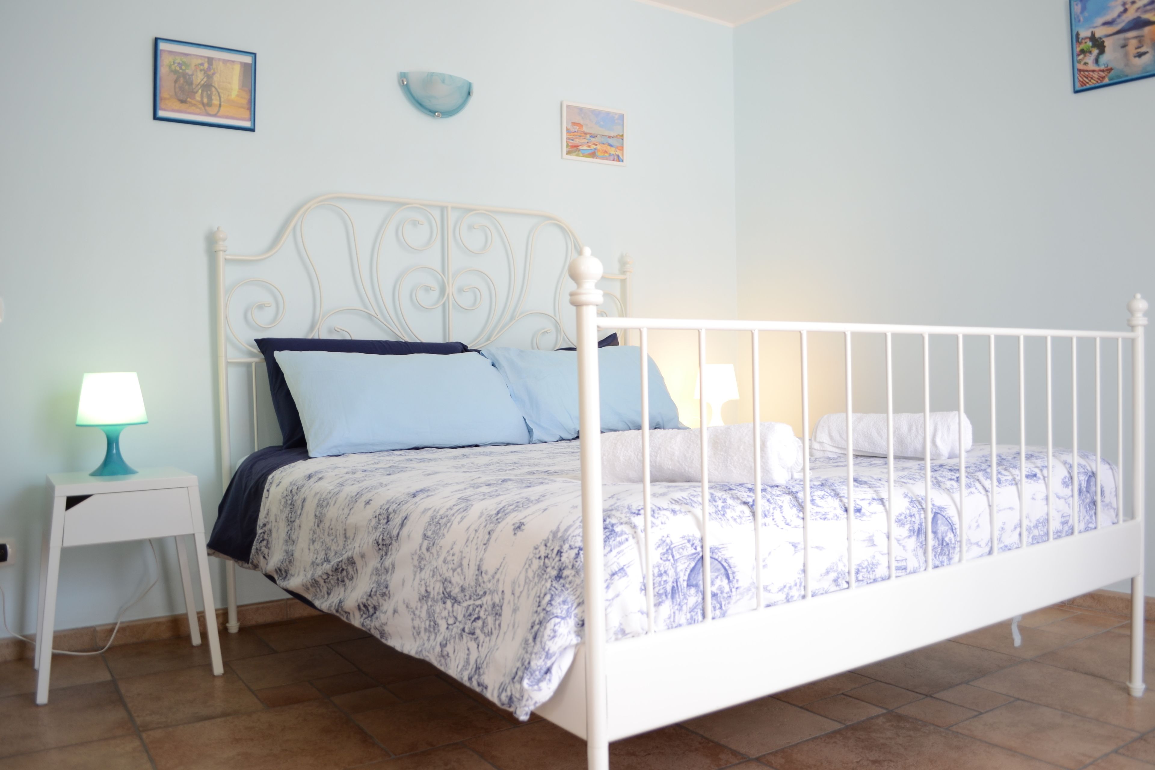 Un dormitorio con un cabecero de metal blanco, una colcha de cuadros azules y blancos, una mesita de noche blanca con una lámpara color turquesa y dos cuadros colgados en la pared.