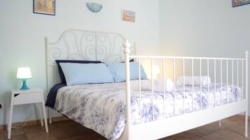 Un dormitorio con un cabecero de metal blanco, una colcha de cuadros azules y blancos, una mesita de noche blanca con una lámpara color turquesa y dos cuadros colgados en la pared.