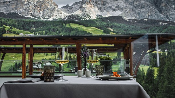Mesa dispuesta para comer con vista a la montaña, una pérgola de madera y una mesa de vidrio.