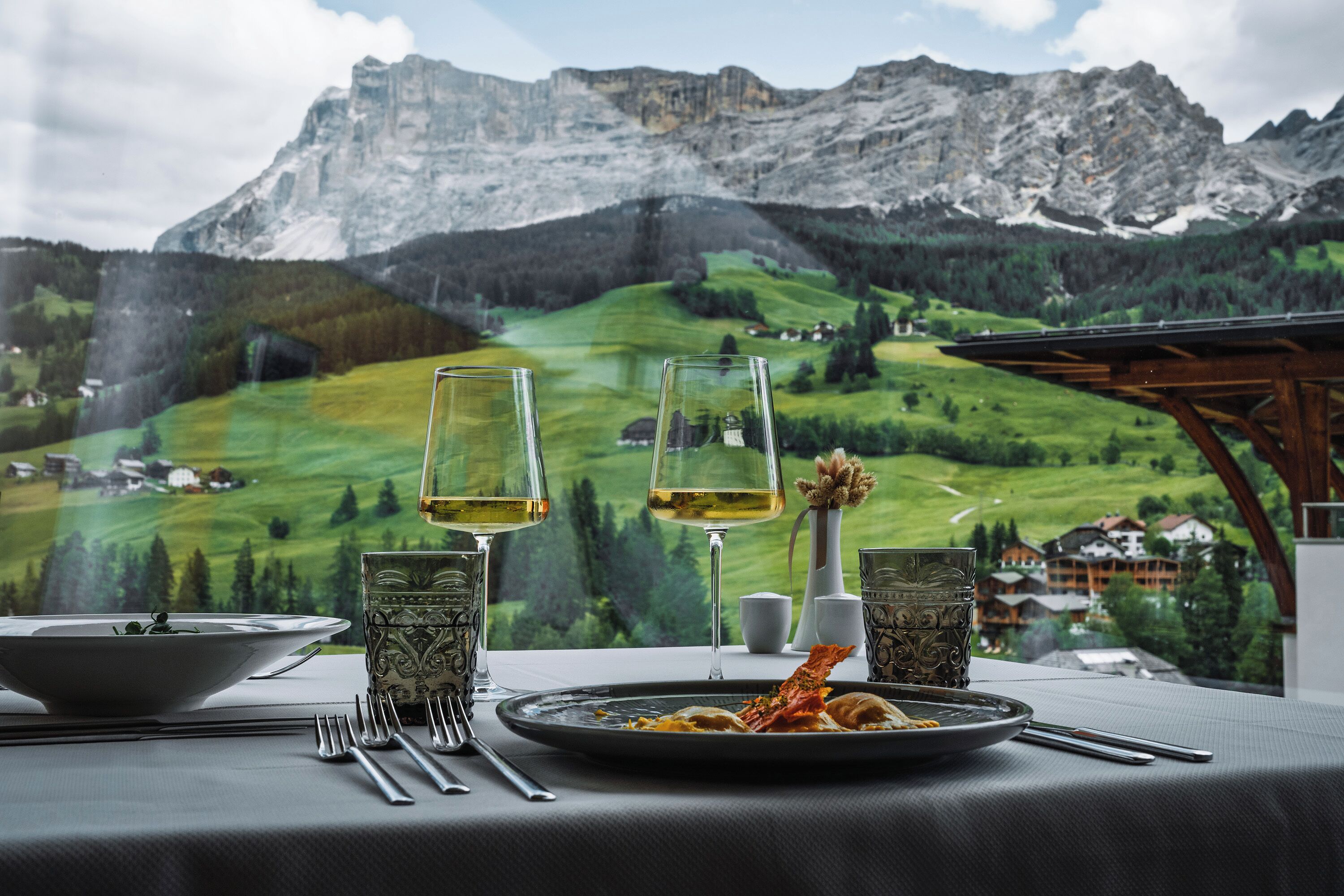 Uma mesa de jantar com vista para uma paisagem montanhosa, dois copos de vinho, um prato de comida e um vaso com uma flor.