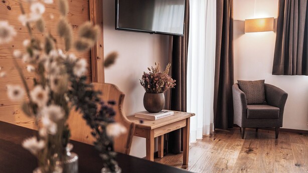 Un salón moderno con suelo de madera, un televisor de pantalla plana montado en la pared, un sillón oscuro, una mesita de madera con un jarrón de flores secas y una lámpara en la mesita auxiliar.