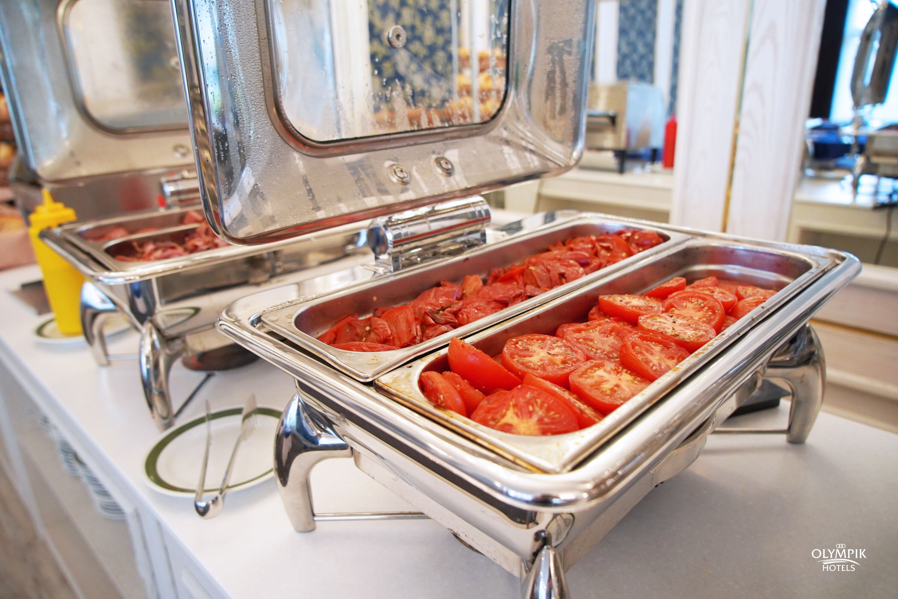 Disposición de bufé con diferentes alimentos, incluyendo tomates en rebanadas, sobre bandejas de acero inoxidable.