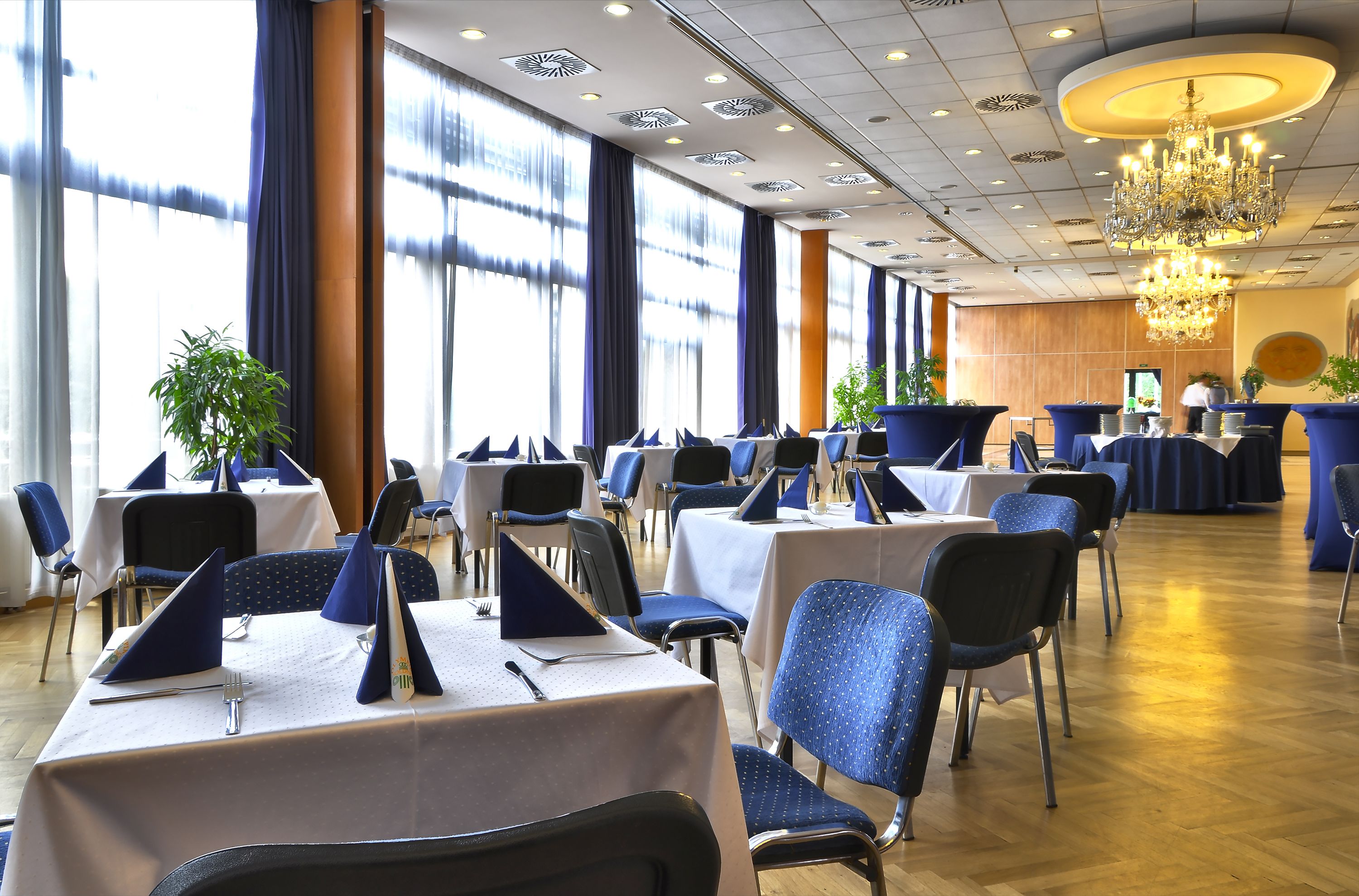 Un salón de conferencias con mesas dispuestas para una reunión, ventanas amplias con cortinas y una araña.
