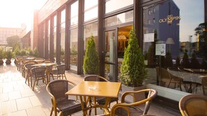 Un espace extérieur aménagé avec des chaises et des tables en osier, un bâtiment doté de grandes fenêtres et une enseigne indiquant l’hôtel Olympiski.