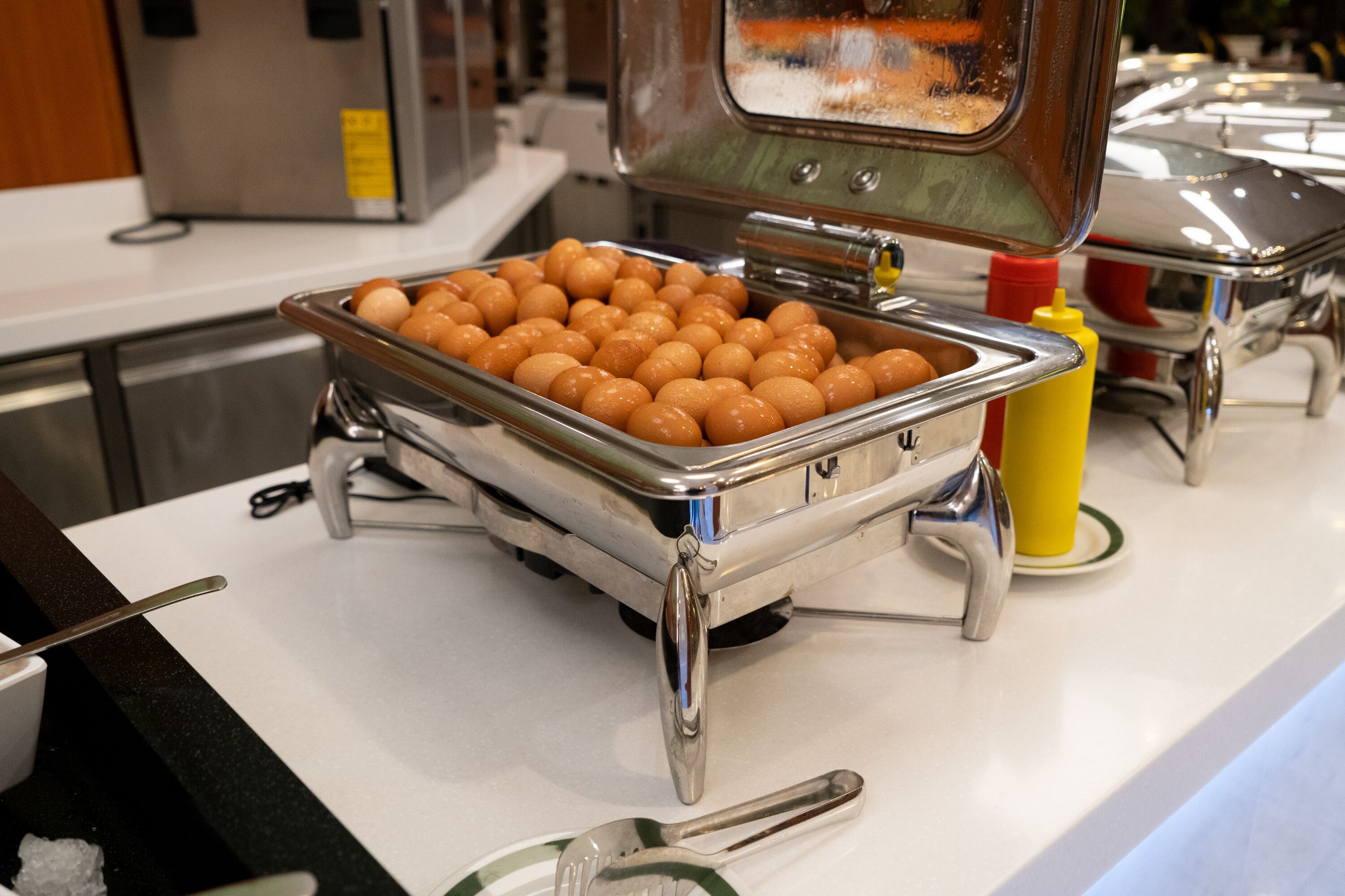 A buffet setup with a tray of round, orange-colored food items, condiment bottles, and a spoon on a white countertop.