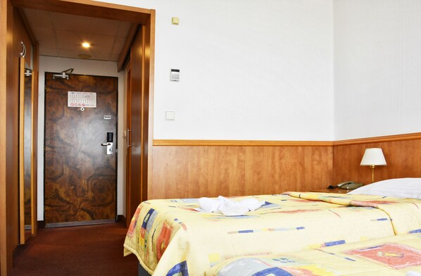 Quarto de hotel com porta de madeira, cama com colcha colorida, abajur e telefone na mesinha de cabeceira.