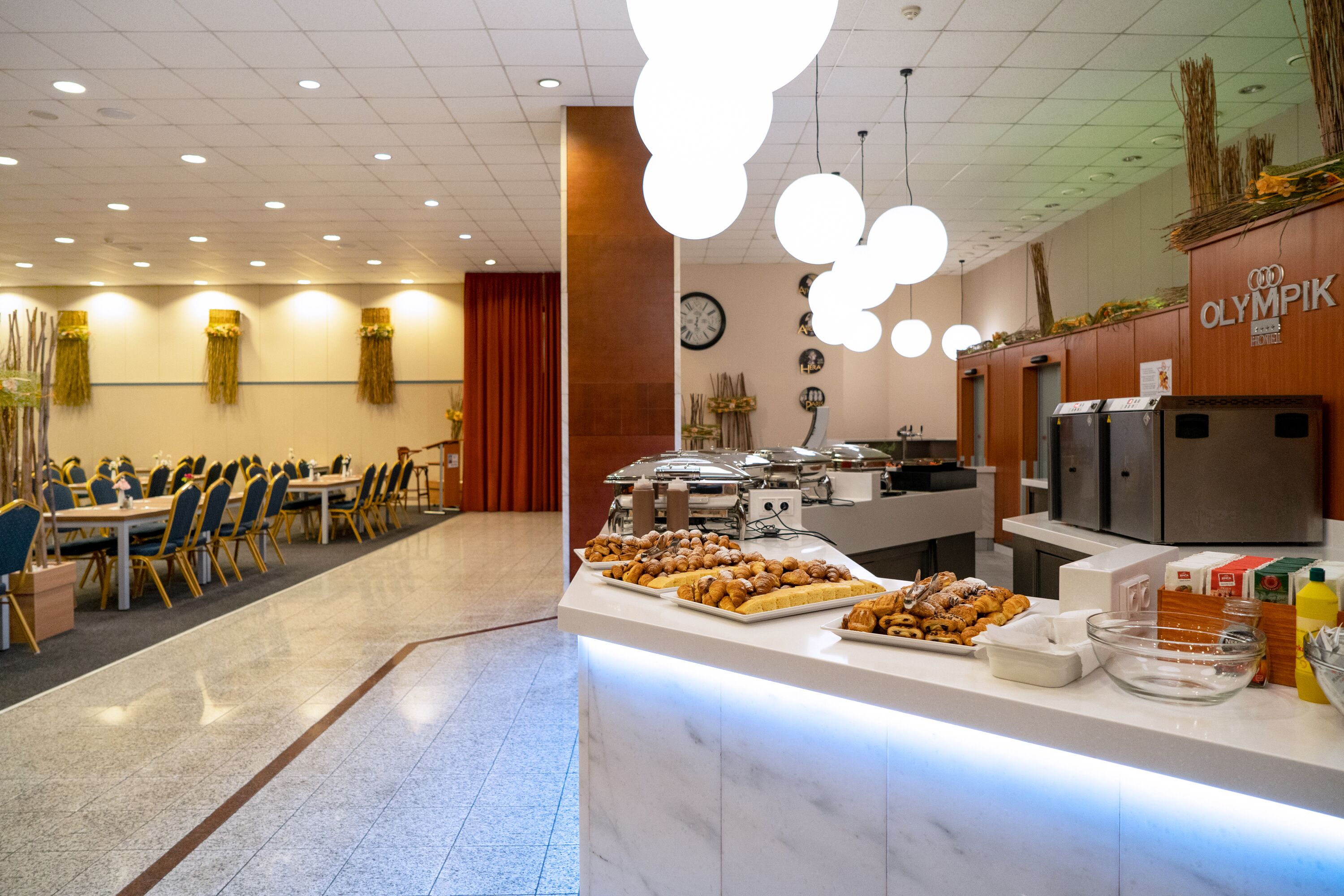 Zona de bufé con diferentes opciones de comida, un reloj en la pared y un letrero para el restaurante Olímpico.
