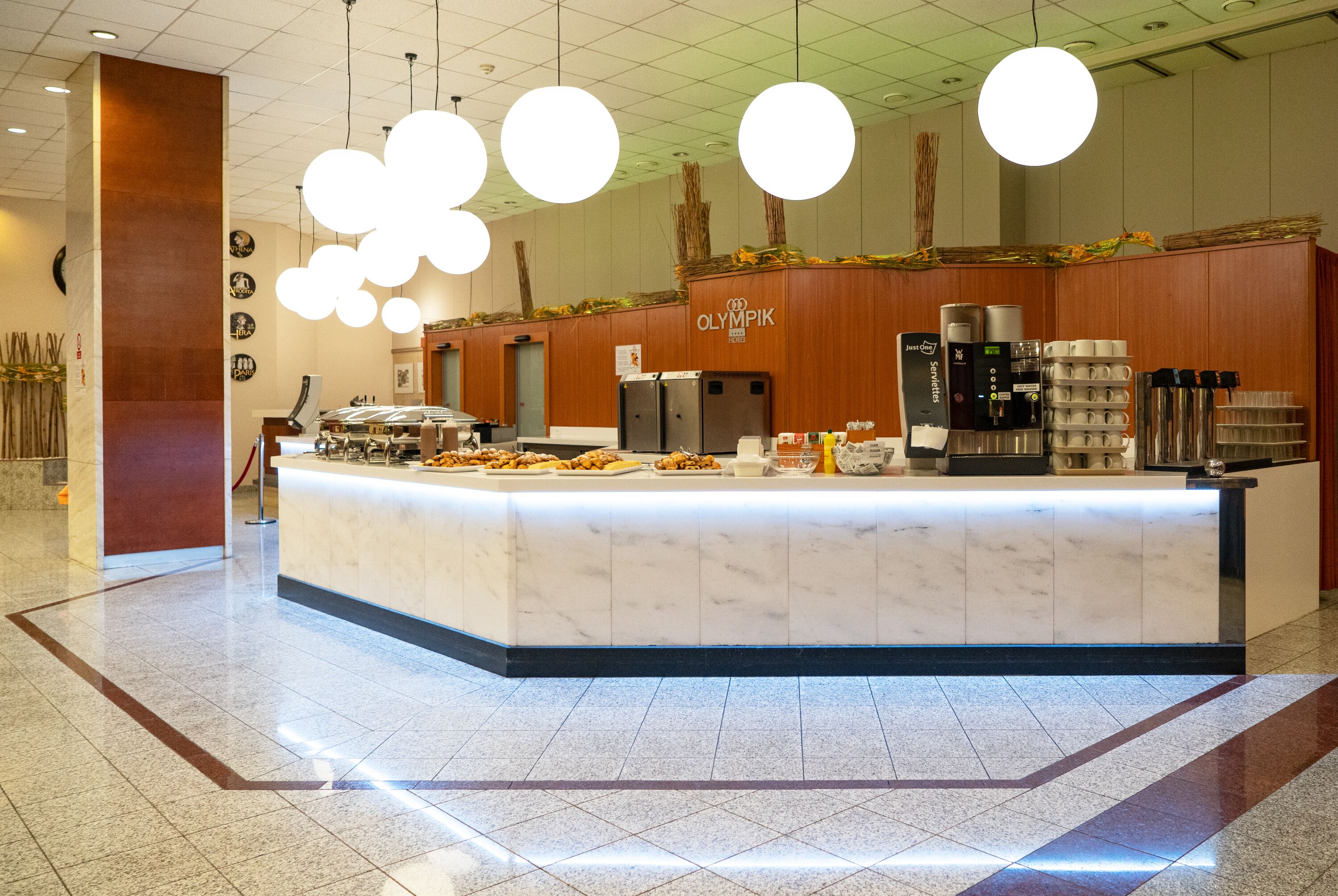 A marble counter with a coffee machine and a refrigerator, surrounded by a wooden wall with the "Olympik" logo.