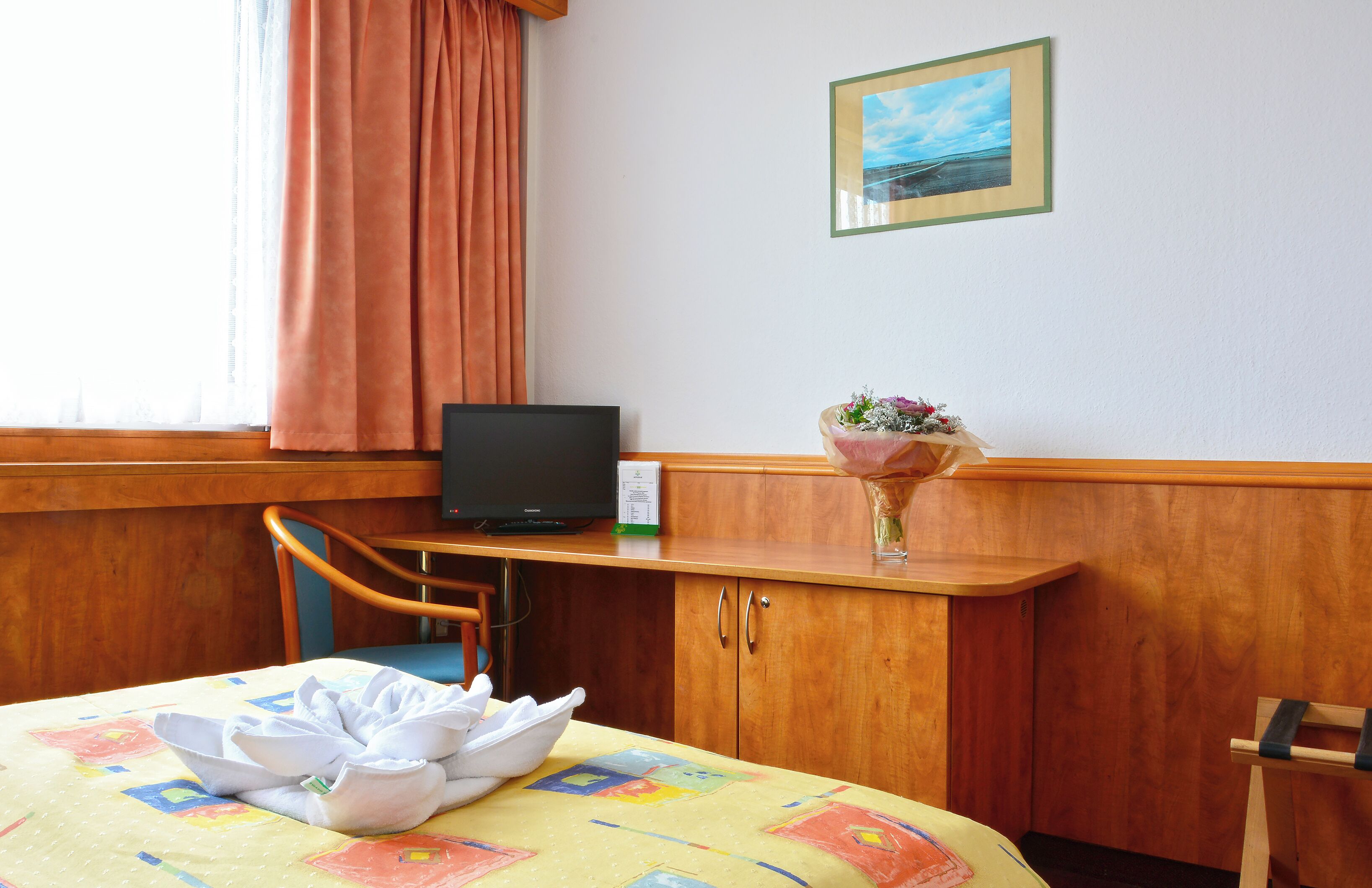 Una habitación de hotel con una cama, un escritorio con televisor, un florero con flores y una ventana con cortinas.
