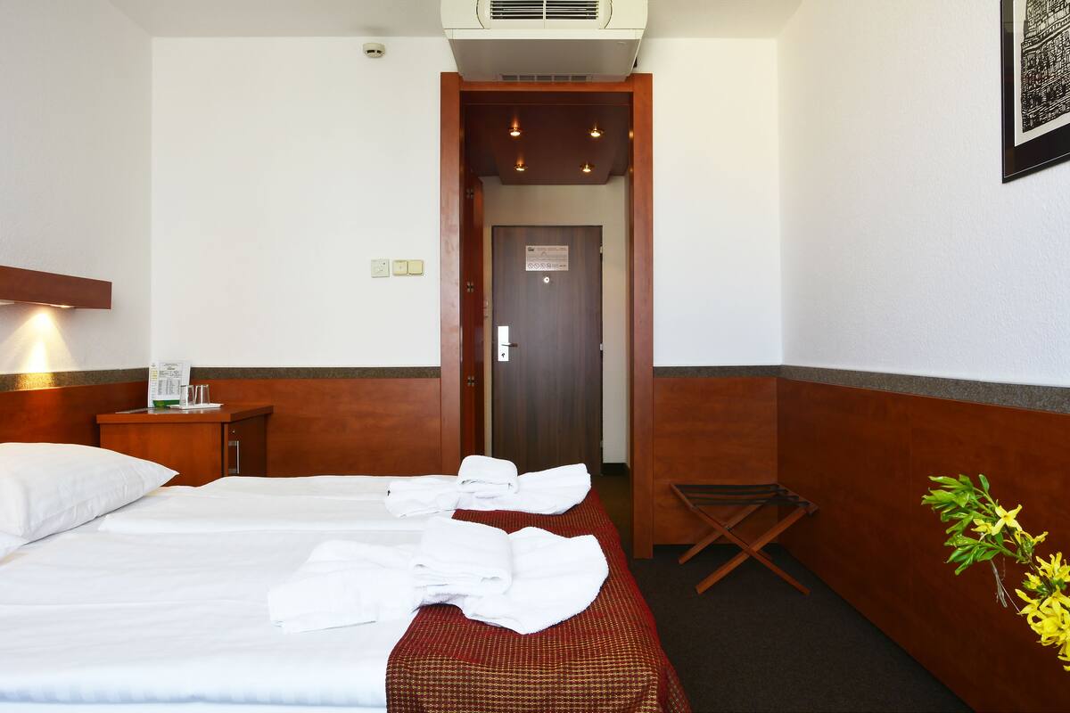 A hotel room with two beds, a wooden headboard, a small table with a lamp, and a door leading to another room.
