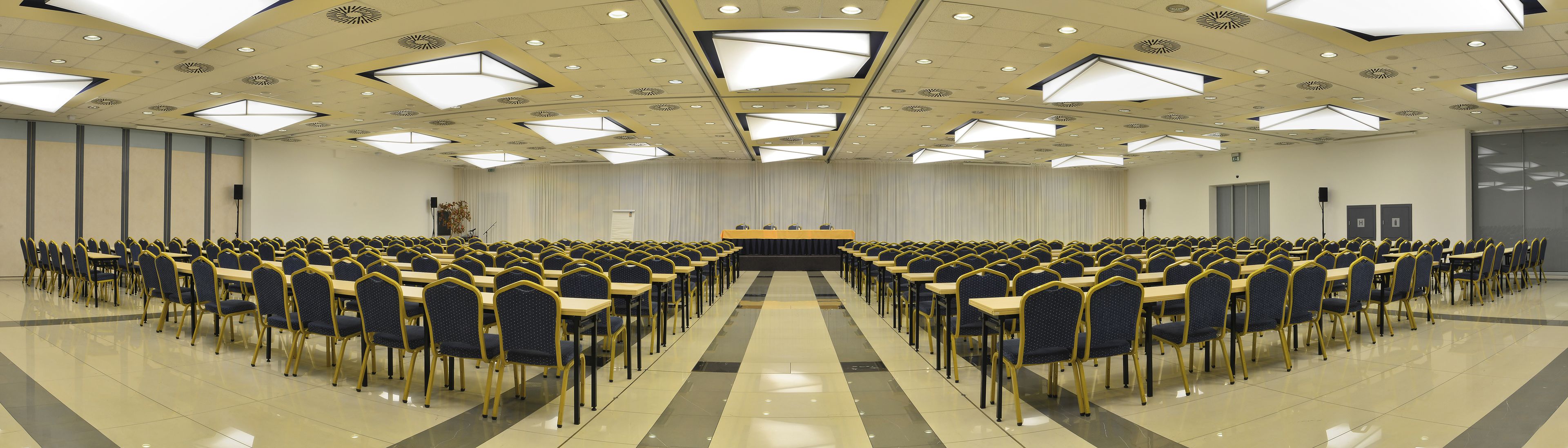 Een congreszaal met rijen tafels en stoelen, een podium aan de voorzijde, en een hoog plafond met geometrische verlichtingsarmaturen.