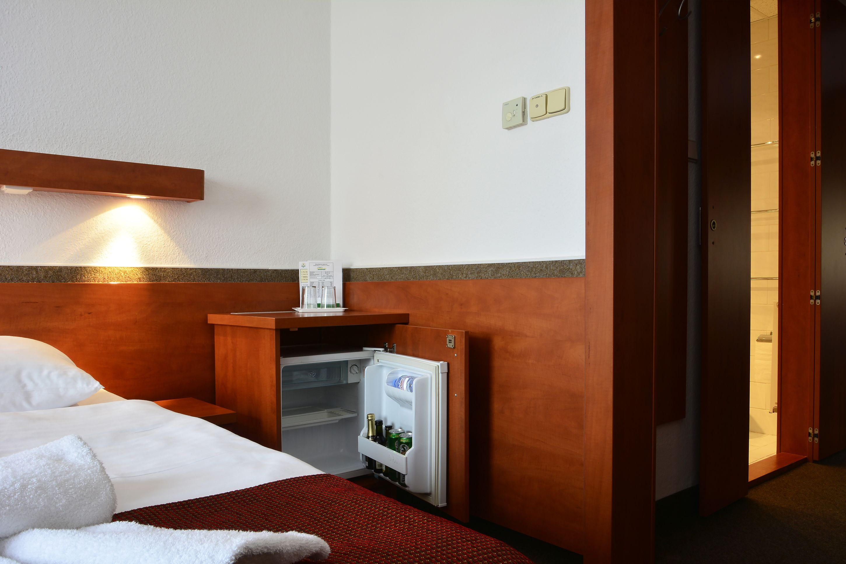 A hotel room with a bed, a small table with a coffee maker, a refrigerator stocked with beverages, and a wooden headboard.